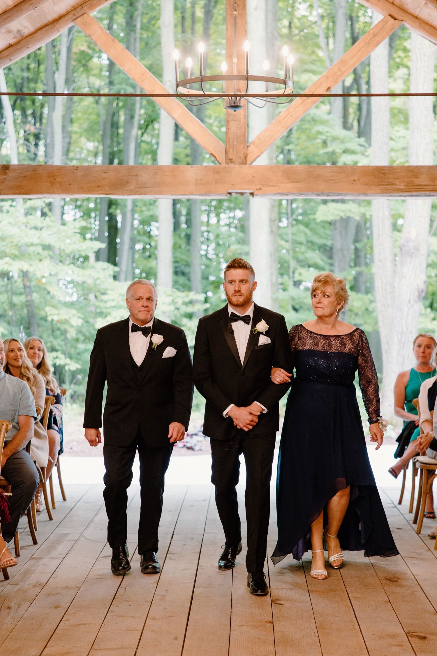 Wedding ceremony processional in rustic wooden pavilion with chandelier and forest backdrop.