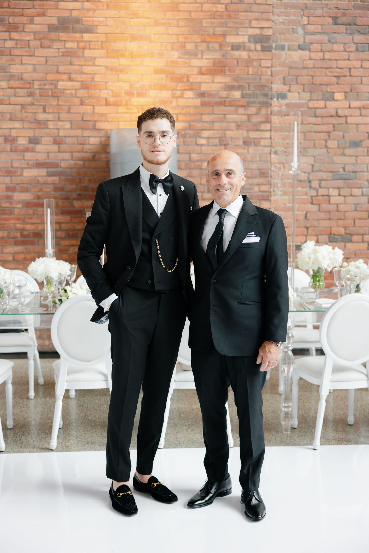 Two people in formal black suits pose against a brick wall at an elegant event venue with white chairs.