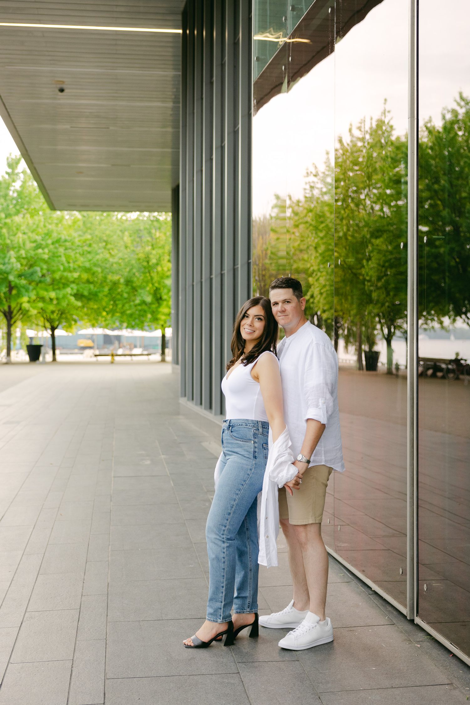 A couple poses casually in a modern outdoor covered walkway with glass panels and greenery in the background.