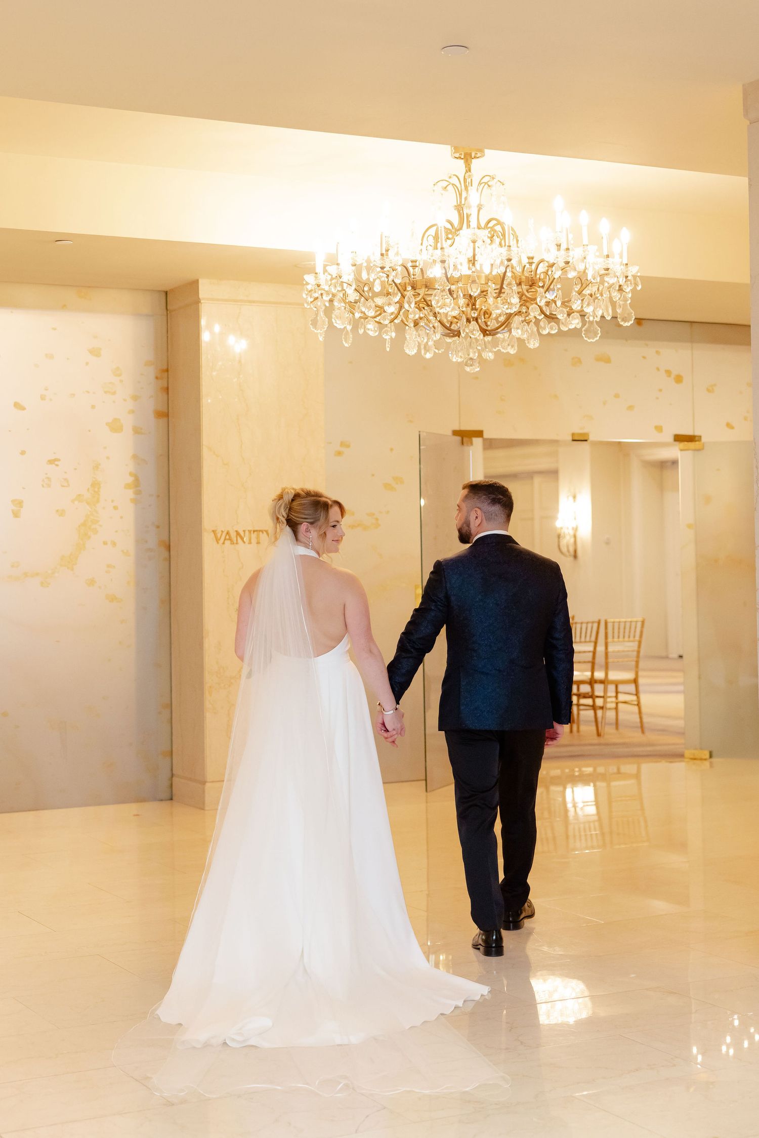 Bride and groom walking together through an elegant white ballroom with ornate lighting fixtures.