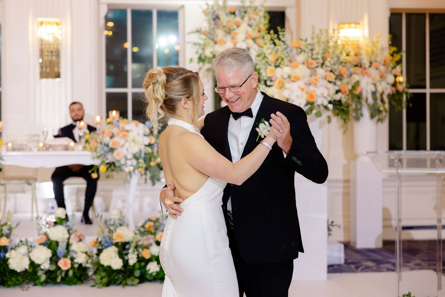 Wedding celebration in an elegant white ballroom with floral decorations and chandeliers.