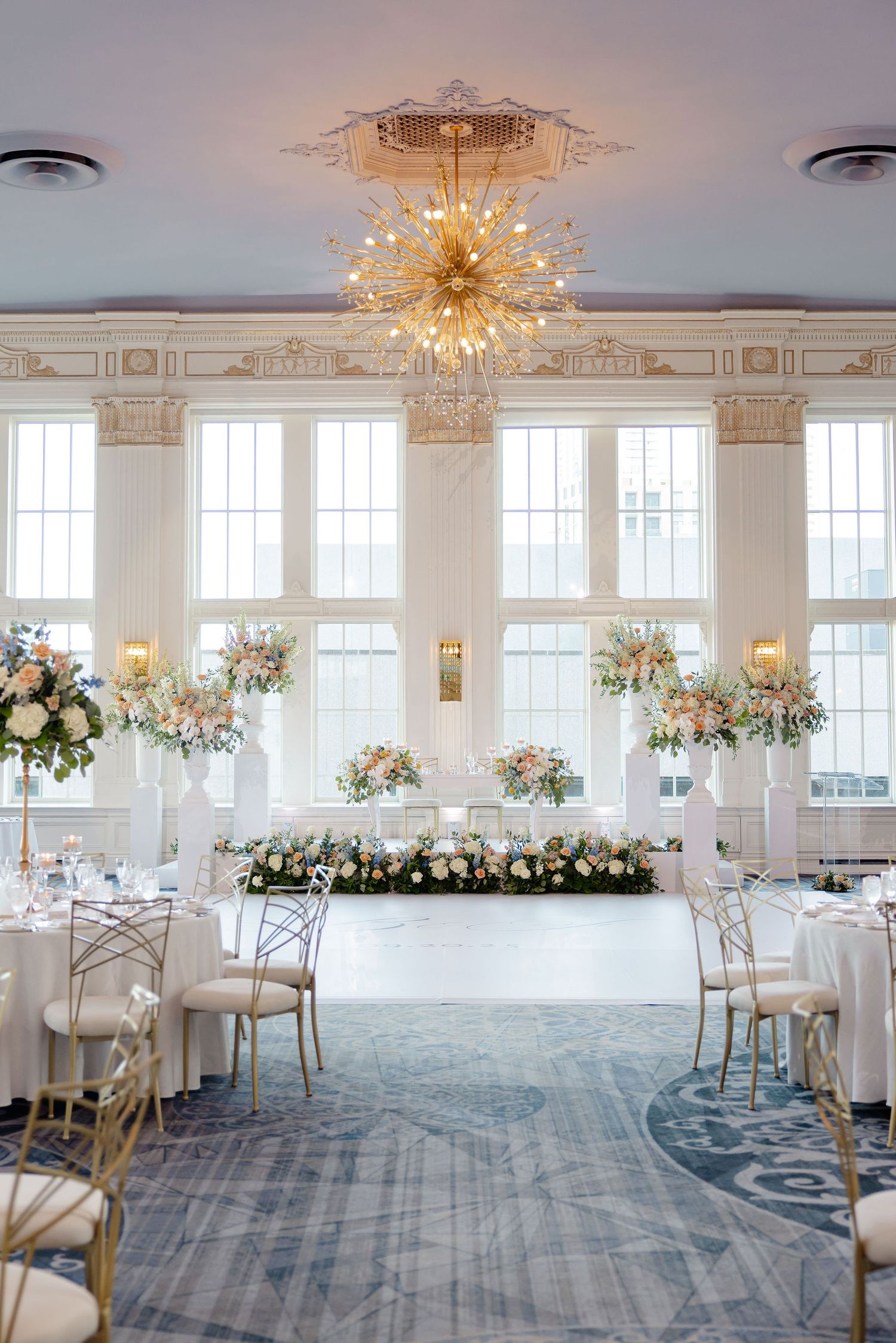 Elegant ballroom with crystal chandeliers, white decor, and blue carpeting set for a luxurious wedding reception.