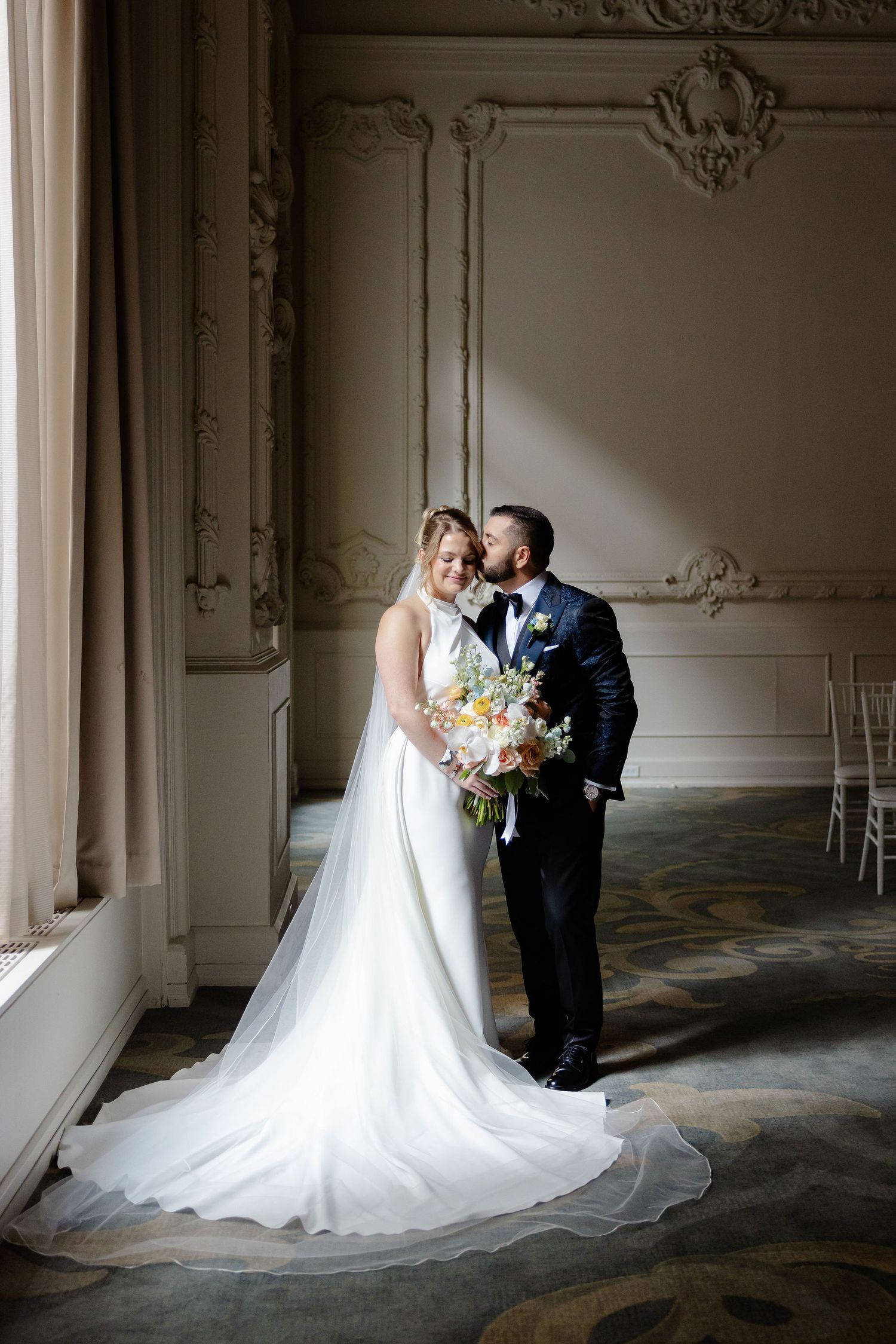Elegant wedding couple embraces in ornate room with white walls and flowing bridal veil.