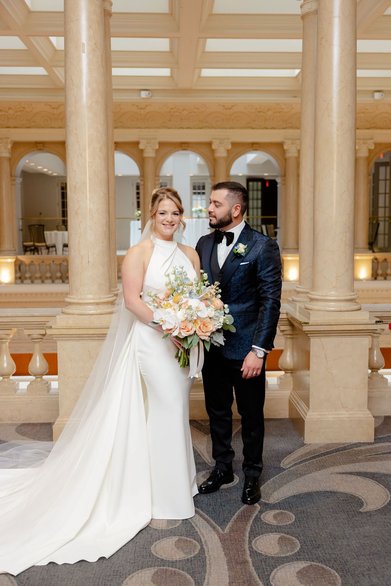 Newlyweds pose in formal attire against classical architecture with columns and ornate ceiling details.