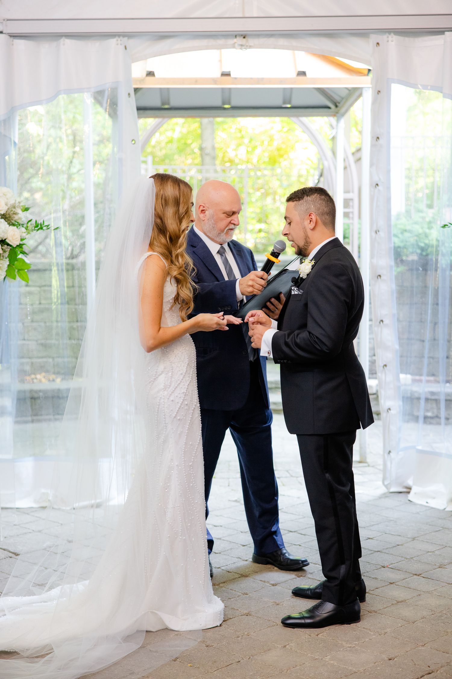 Wedding ceremony moment showing exchange of rings under white tent with natural backdrop.