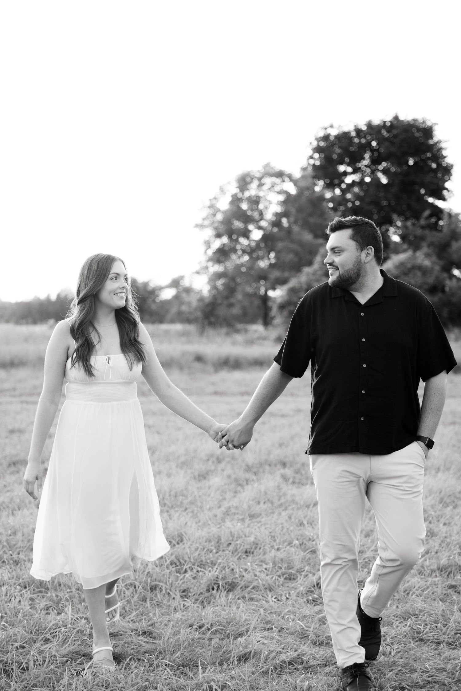 A black and white photo captures a couple walking hand-in-hand through a grassy field at sunset.