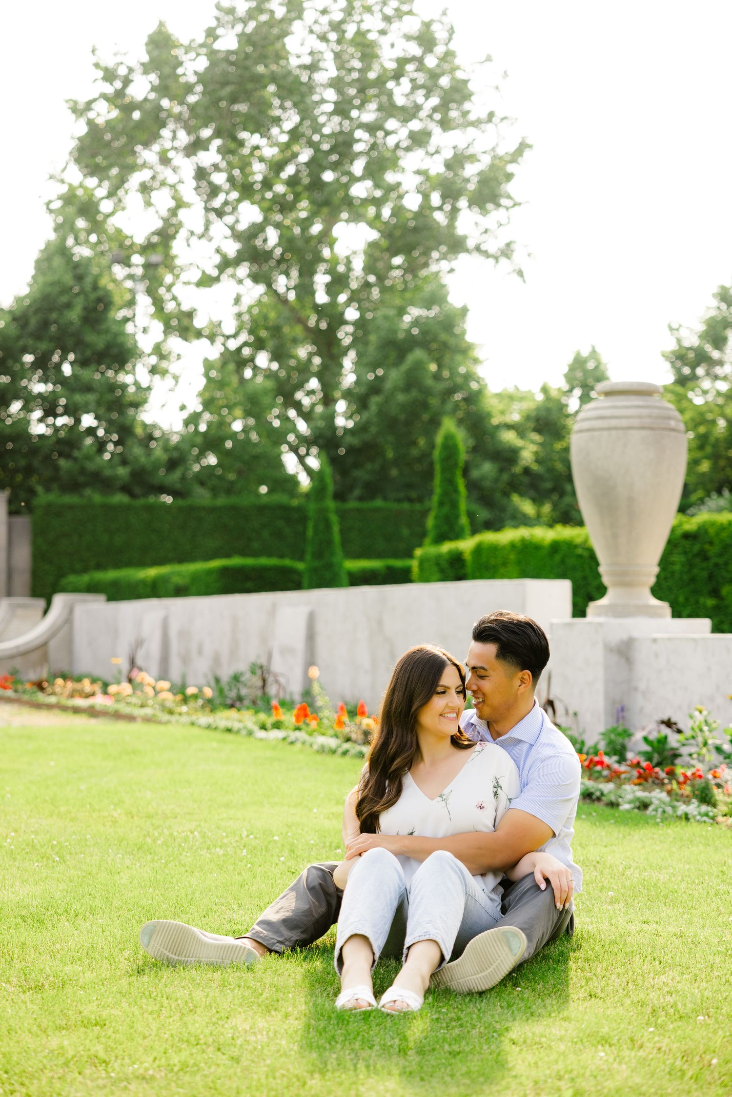 A couple sits together on manicured lawn near ornamental urns and garden flowers.