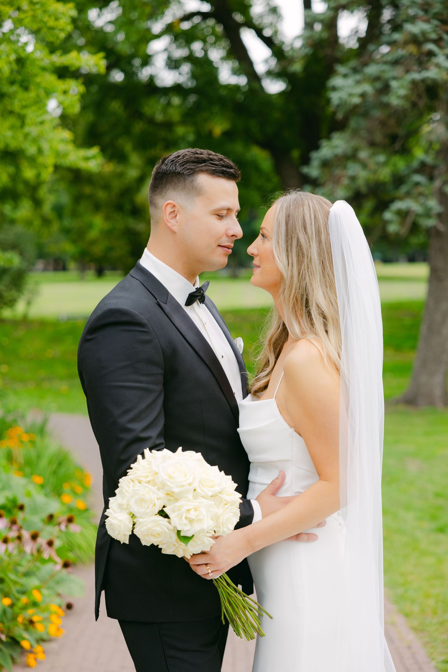 A wedding couple shares a tender moment while holding a white rose bouquet in a garden.