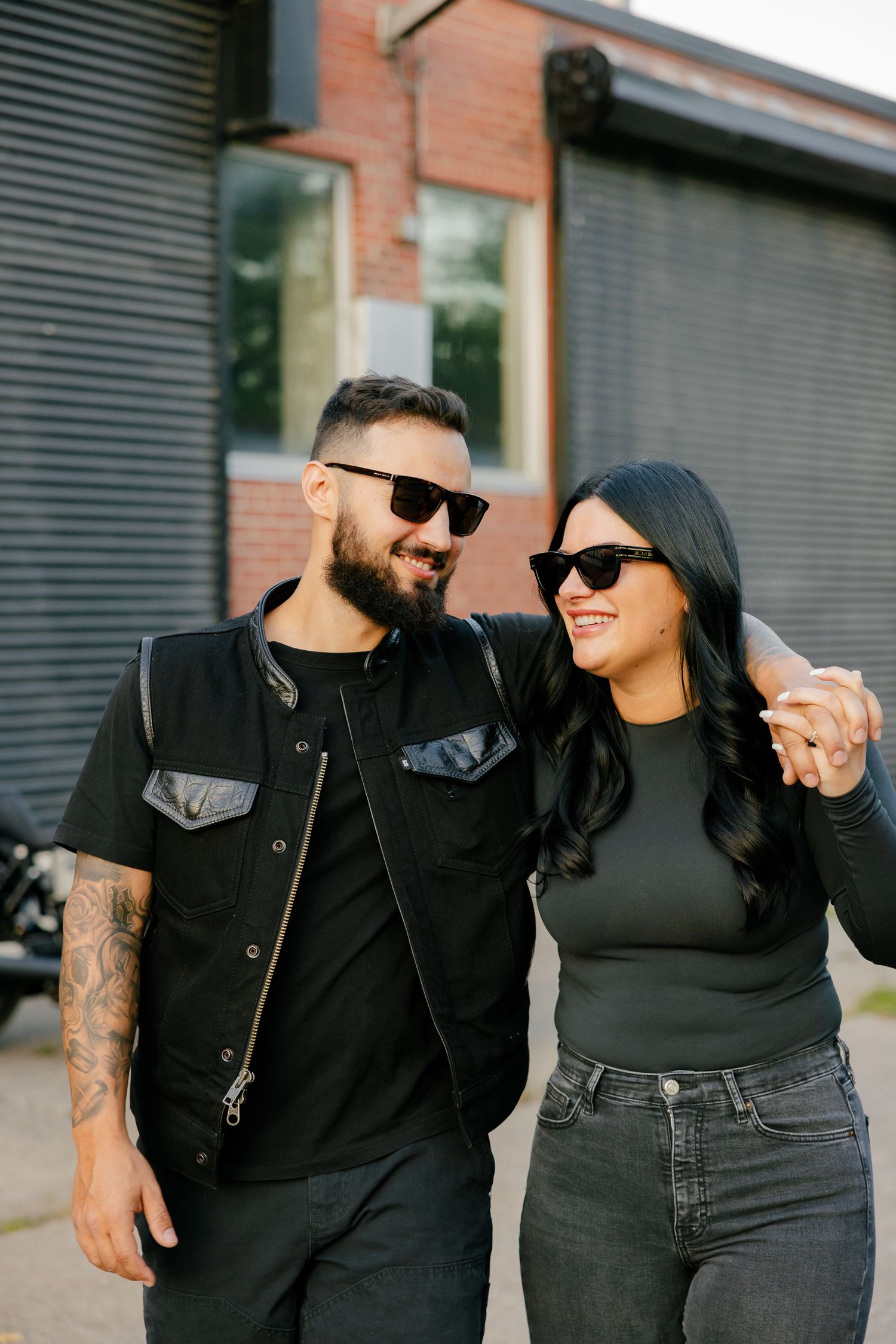 A couple in coordinated black outfits smiling at each other against a brick and metal wall.
