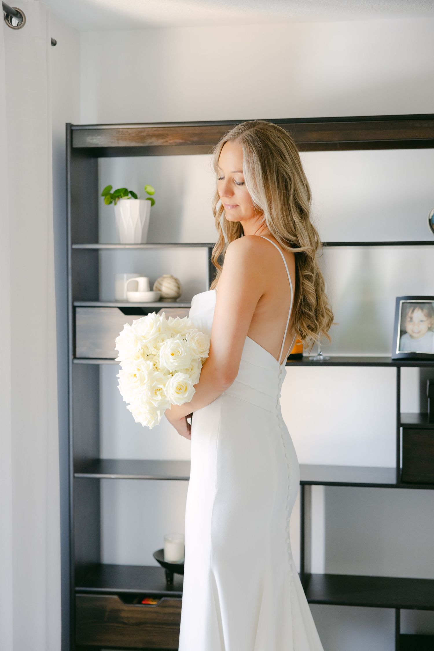 Elegant bride in fitted white gown holds a bouquet of white flowers against modern shelving.