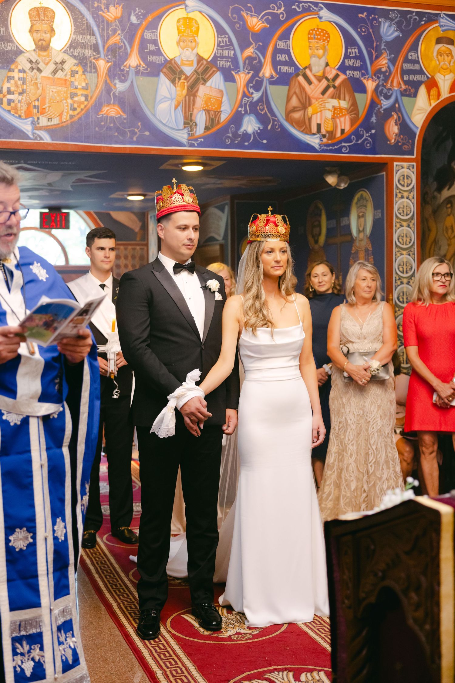 Orthodox wedding ceremony with priest in blue robes leading service against backdrop of religious icons.