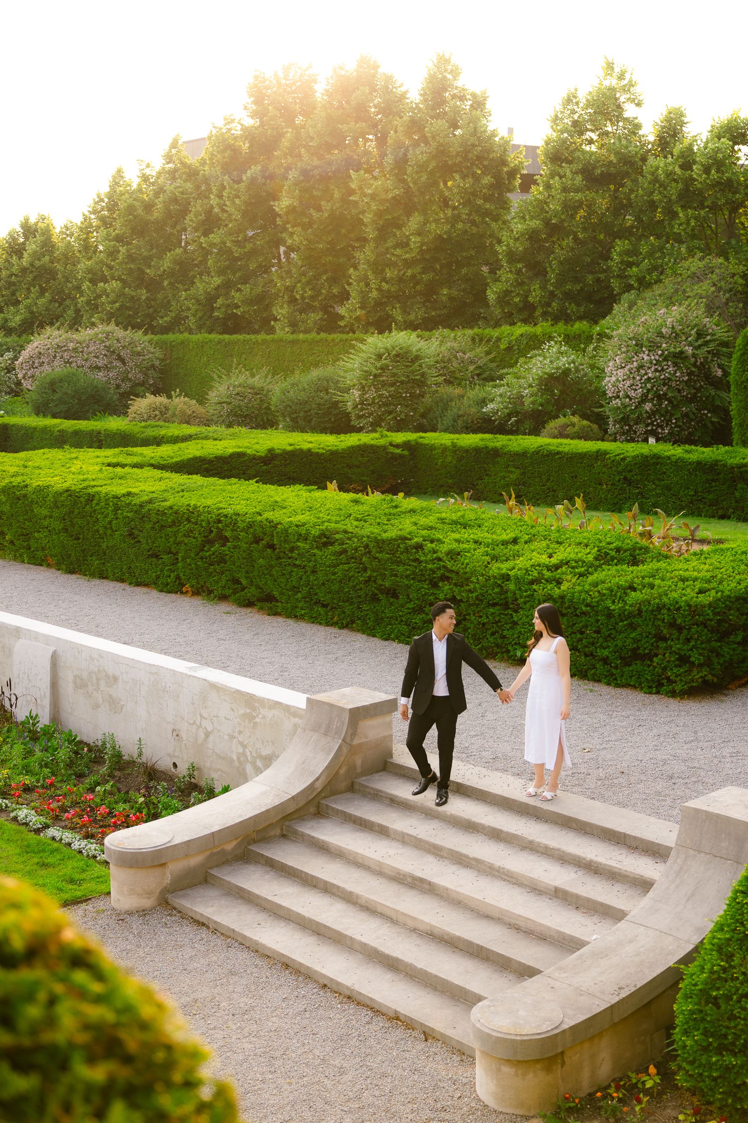 A couple holding hands while standing on curved stone steps in a sunlit formal garden.