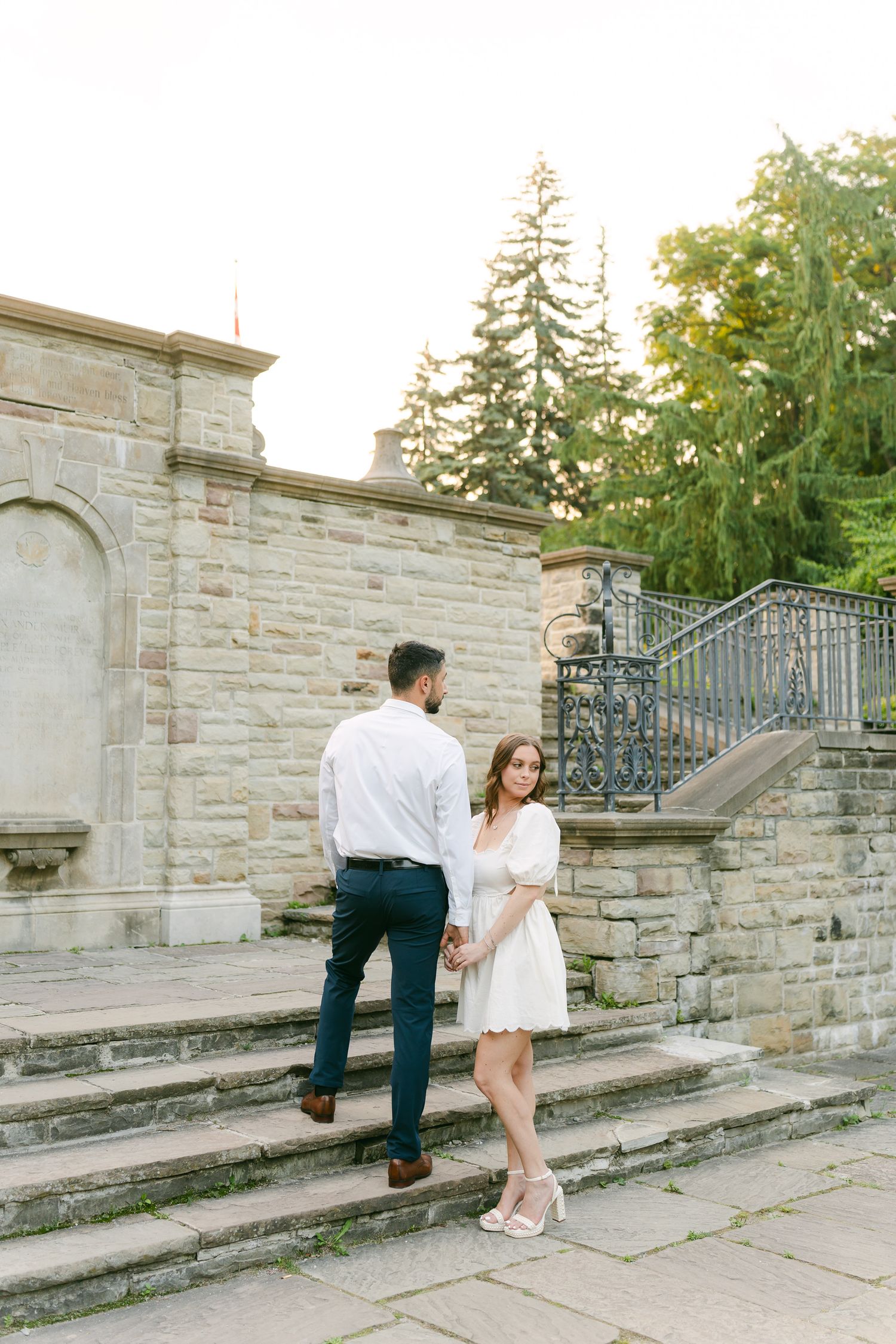 A couple shares a tender moment on stone steps with decorative iron railings and evergreen trees.