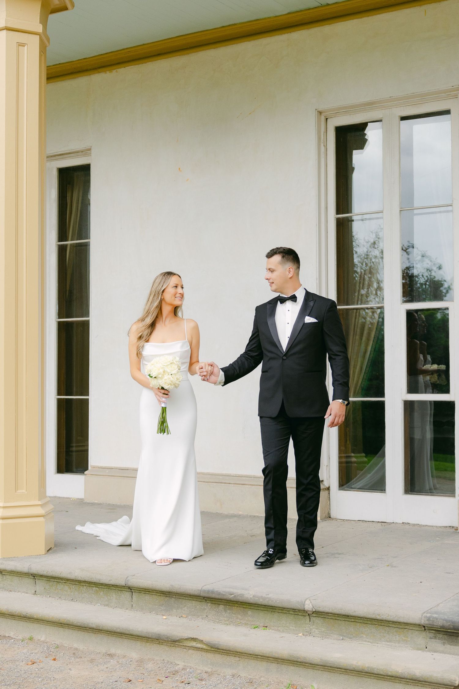 Bride holding bouquet while walking with groom on classic stone porch.