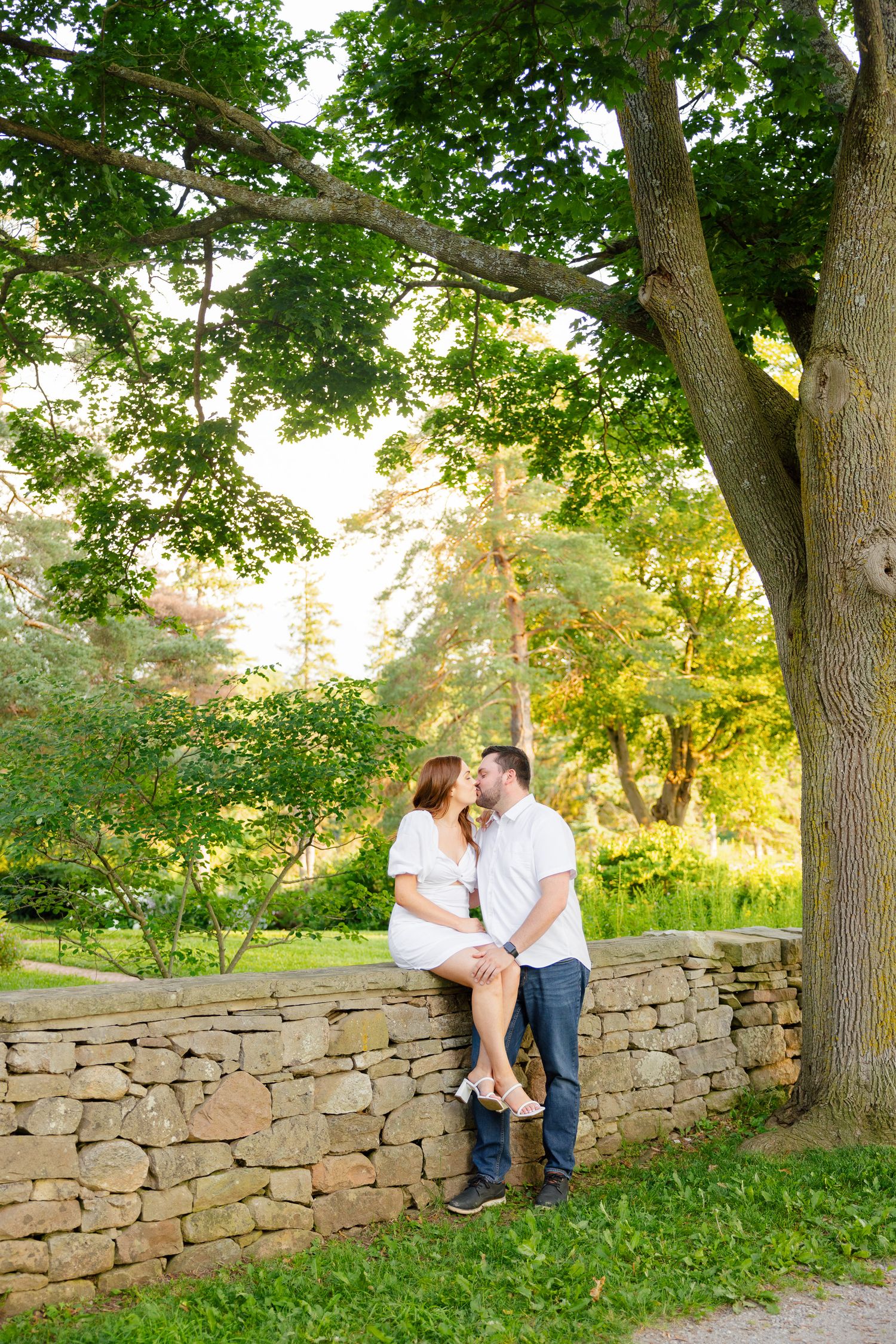 A tender moment between a couple sitting on a stone wall surrounded by lush summer foliage.