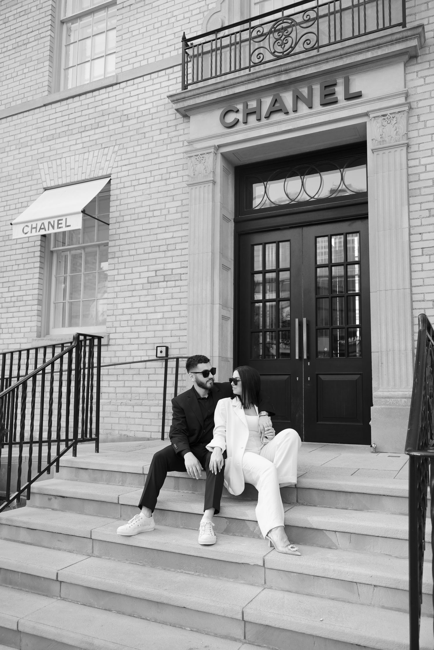 A stylish couple sits on steps outside a Chanel boutique storefront in black and white photography.