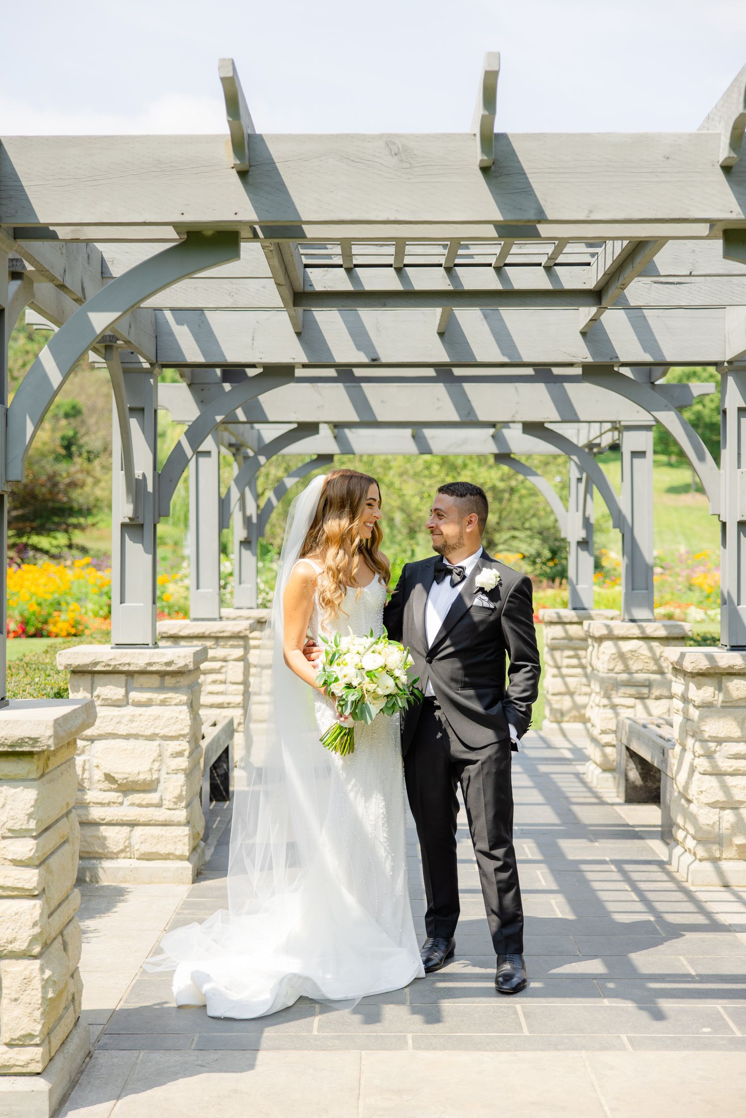 Newlyweds share a tender moment under an elegant stone and wood pergola in a garden setting.