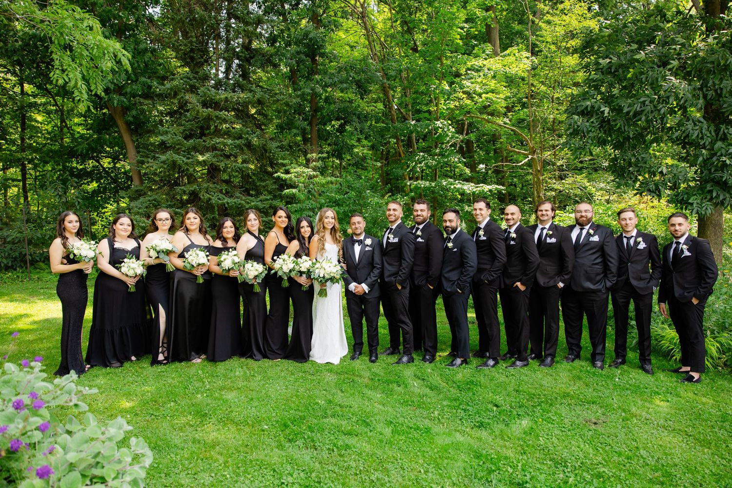 Large wedding party in black attire poses on grass with trees in background.