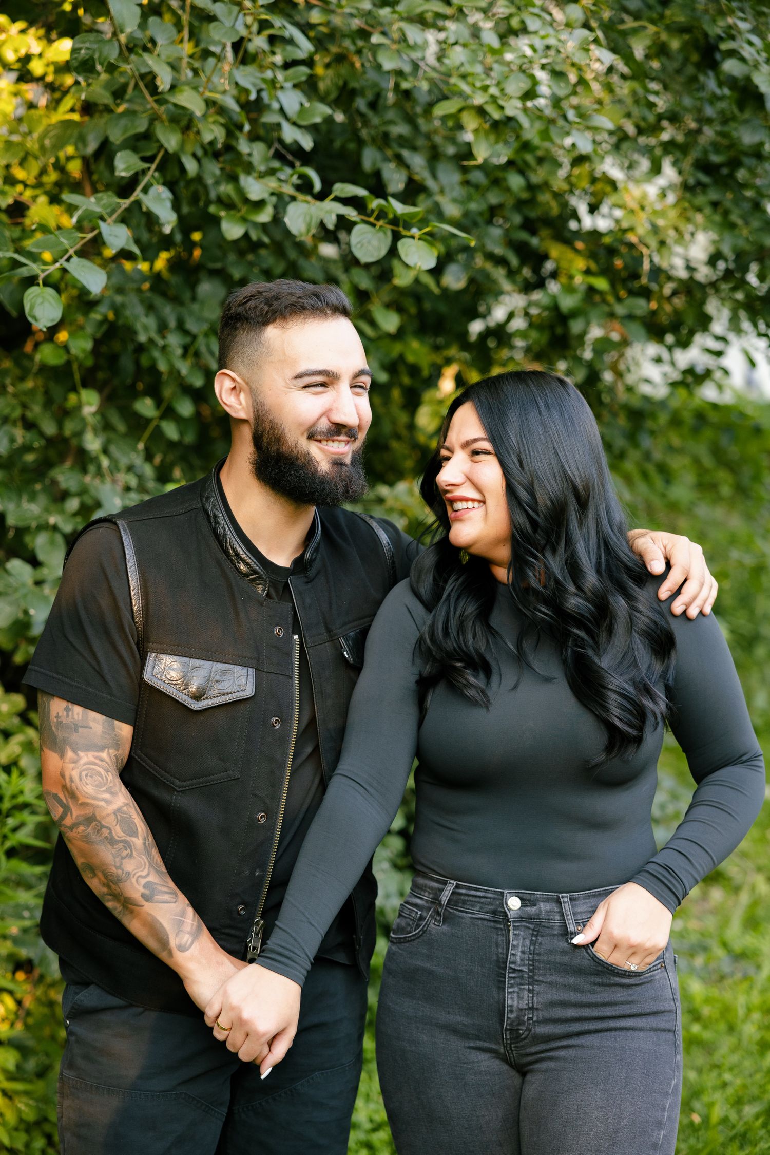 A couple laughs together while posing for a photo in matching dark outfits outdoors.