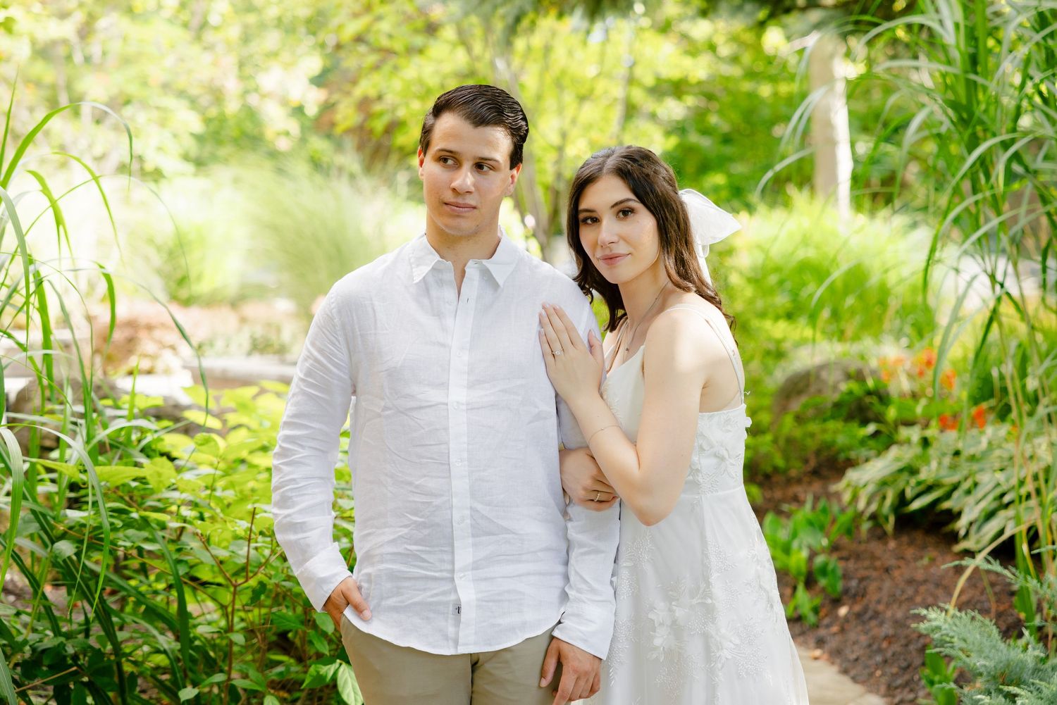 A couple poses together in a sunlit garden wearing coordinating white outfits.