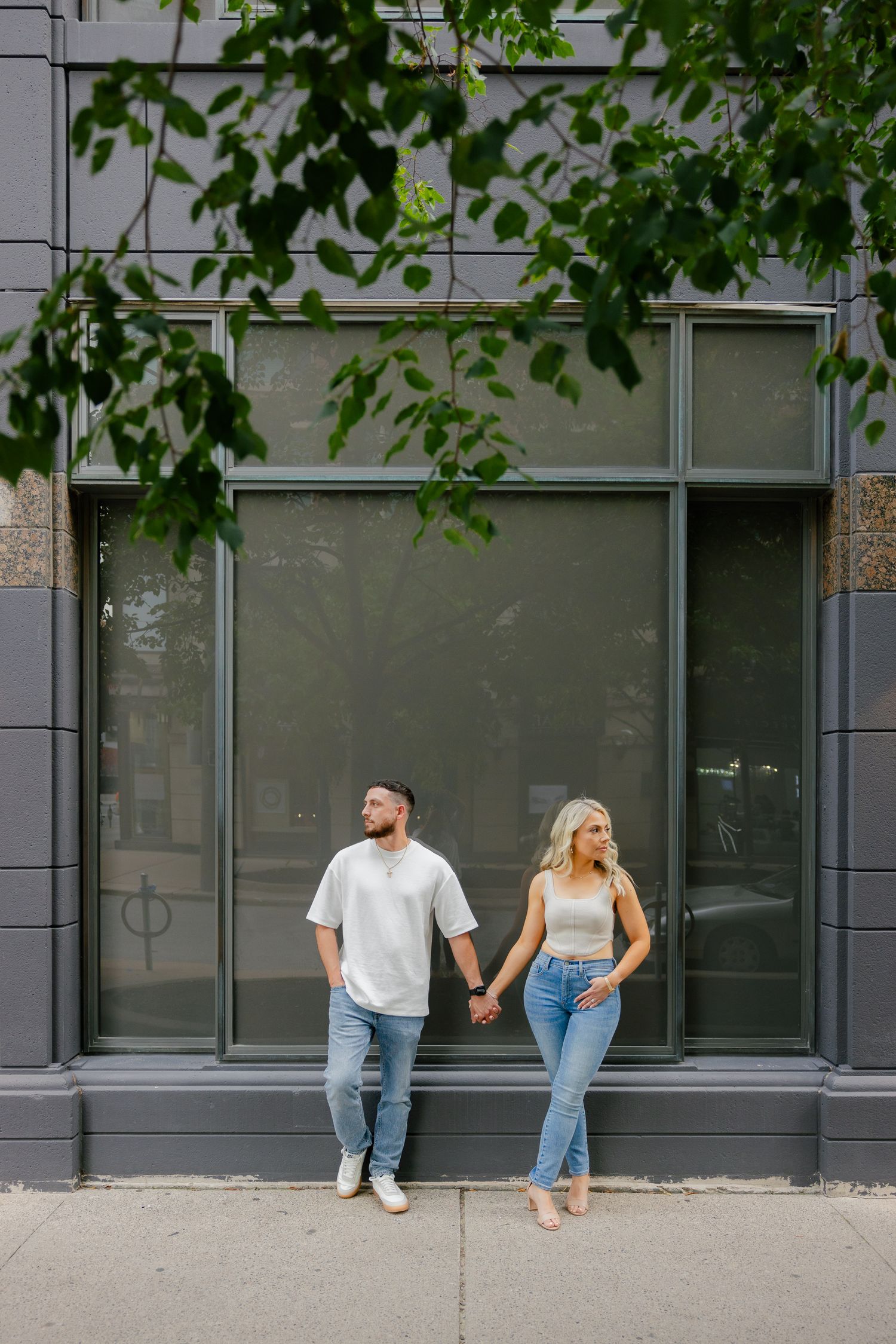 Stylish pair in casual wear holding hands against modern building exterior.