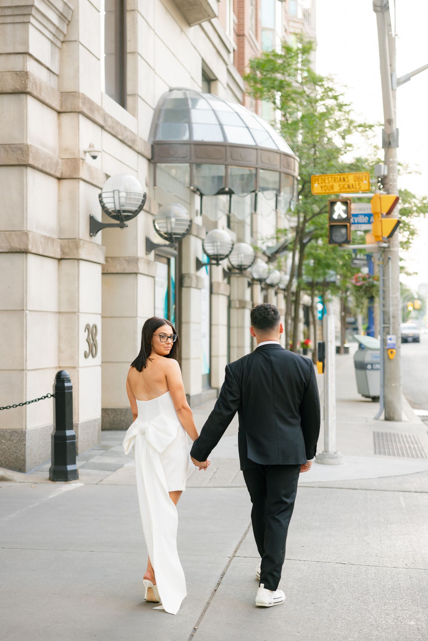 Elegant couple walking down a city street with distinctive architectural features and sunlit backdrop.
