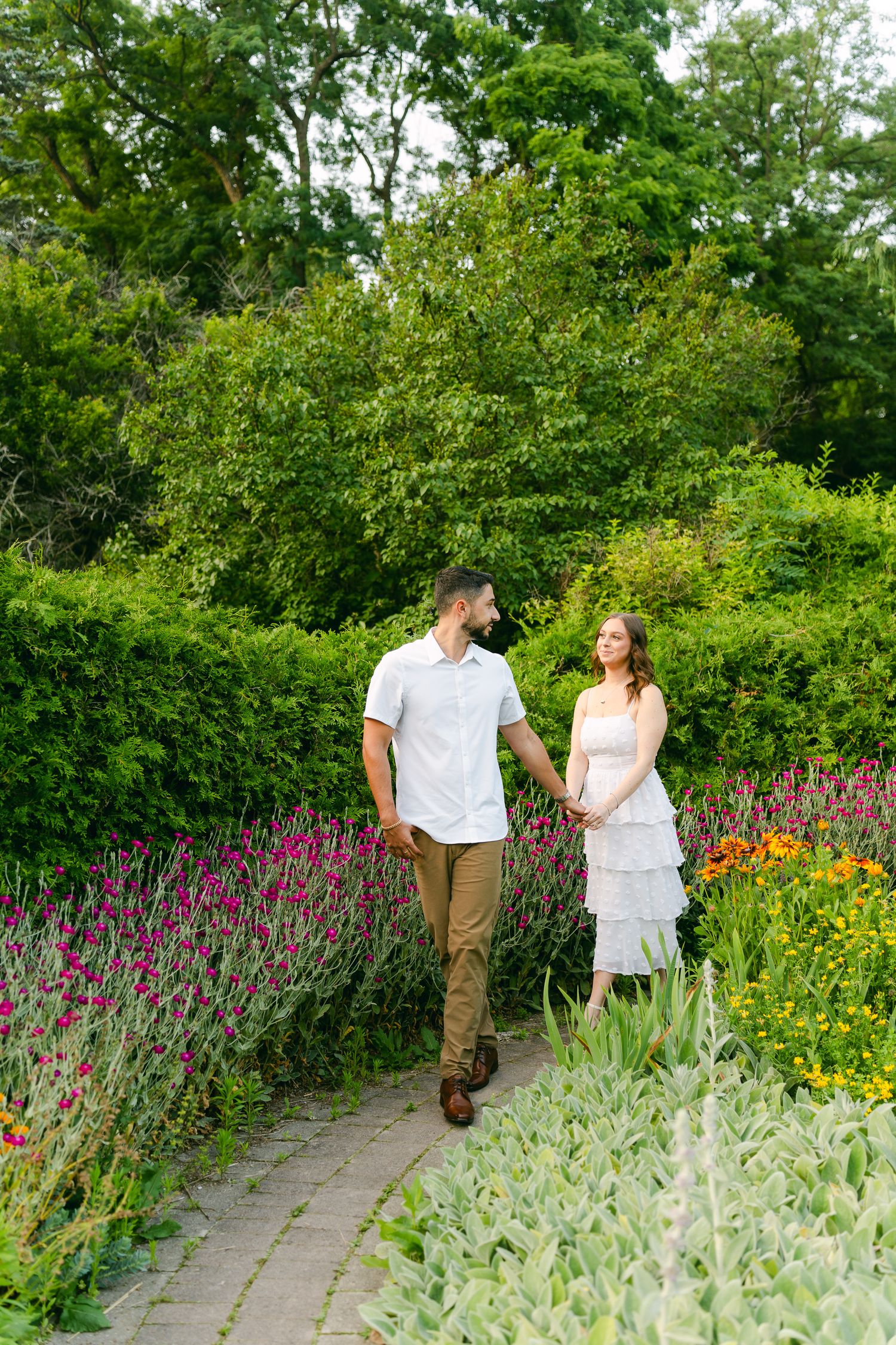 Couple walking hand in hand along a garden path with vibrant purple flowers and lush greenery.