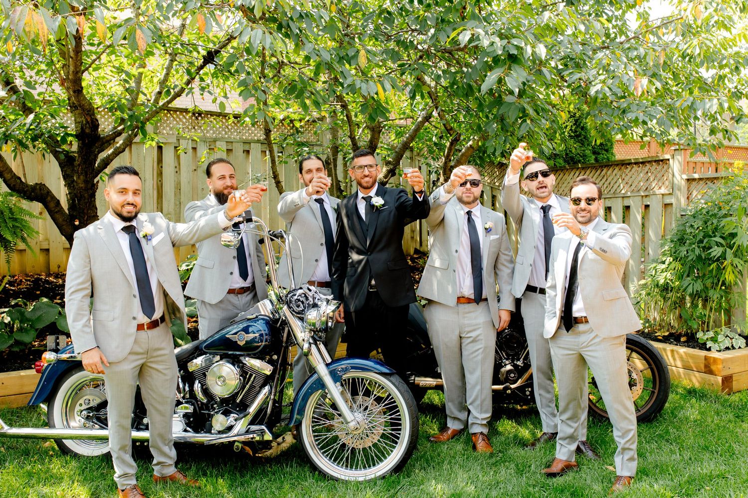 Wedding party celebrating with drinks around vintage motorcycles in a backyard.