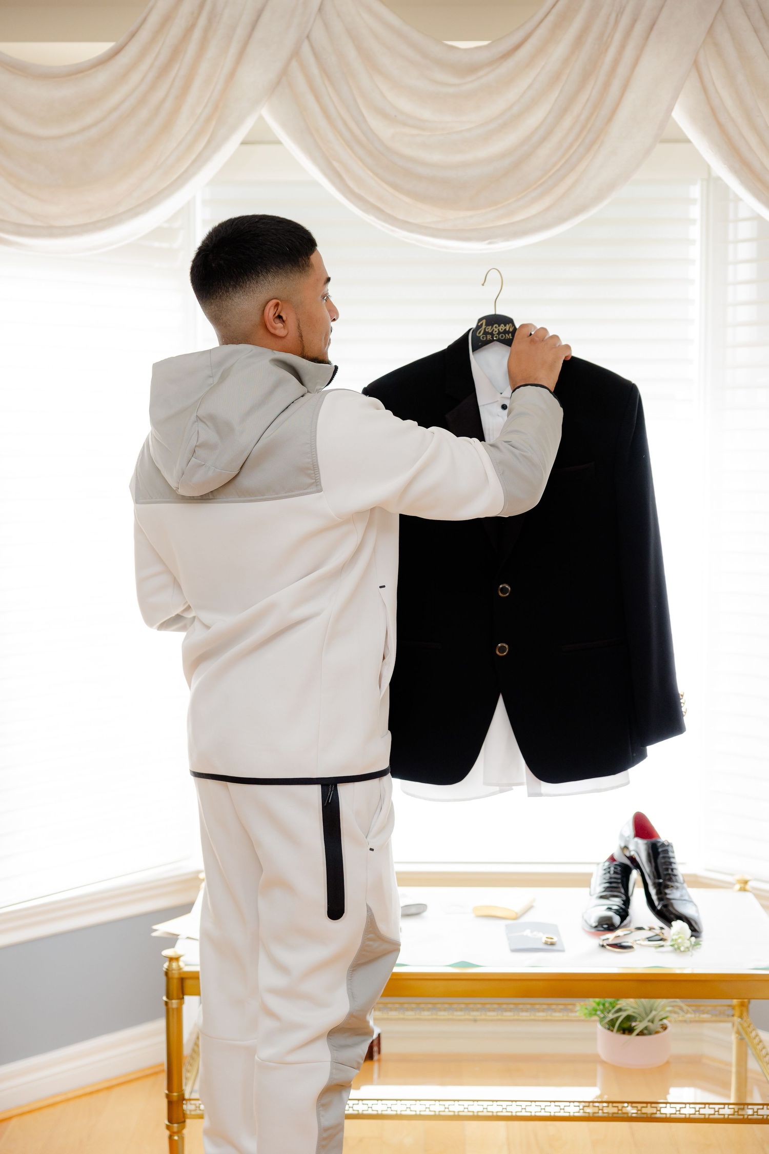 Someone in white attire holding up a formal black suit jacket against draped window.