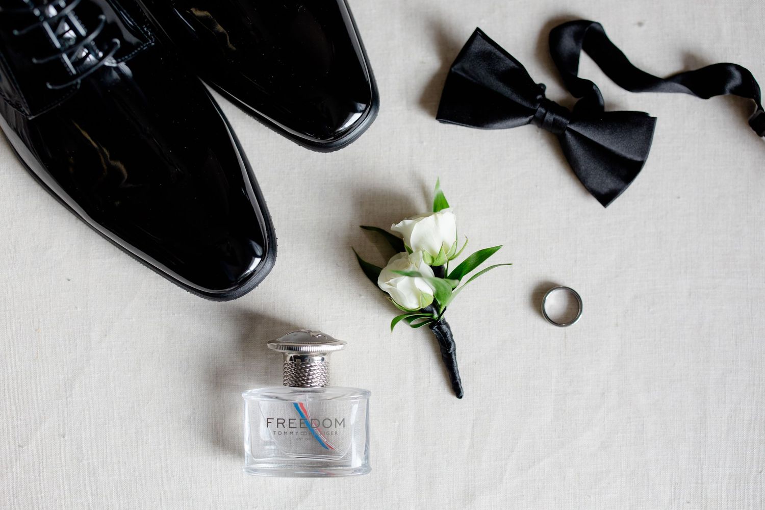Wedding accessories featuring patent leather shoes, white boutonniere, bow tie and cologne bottle.