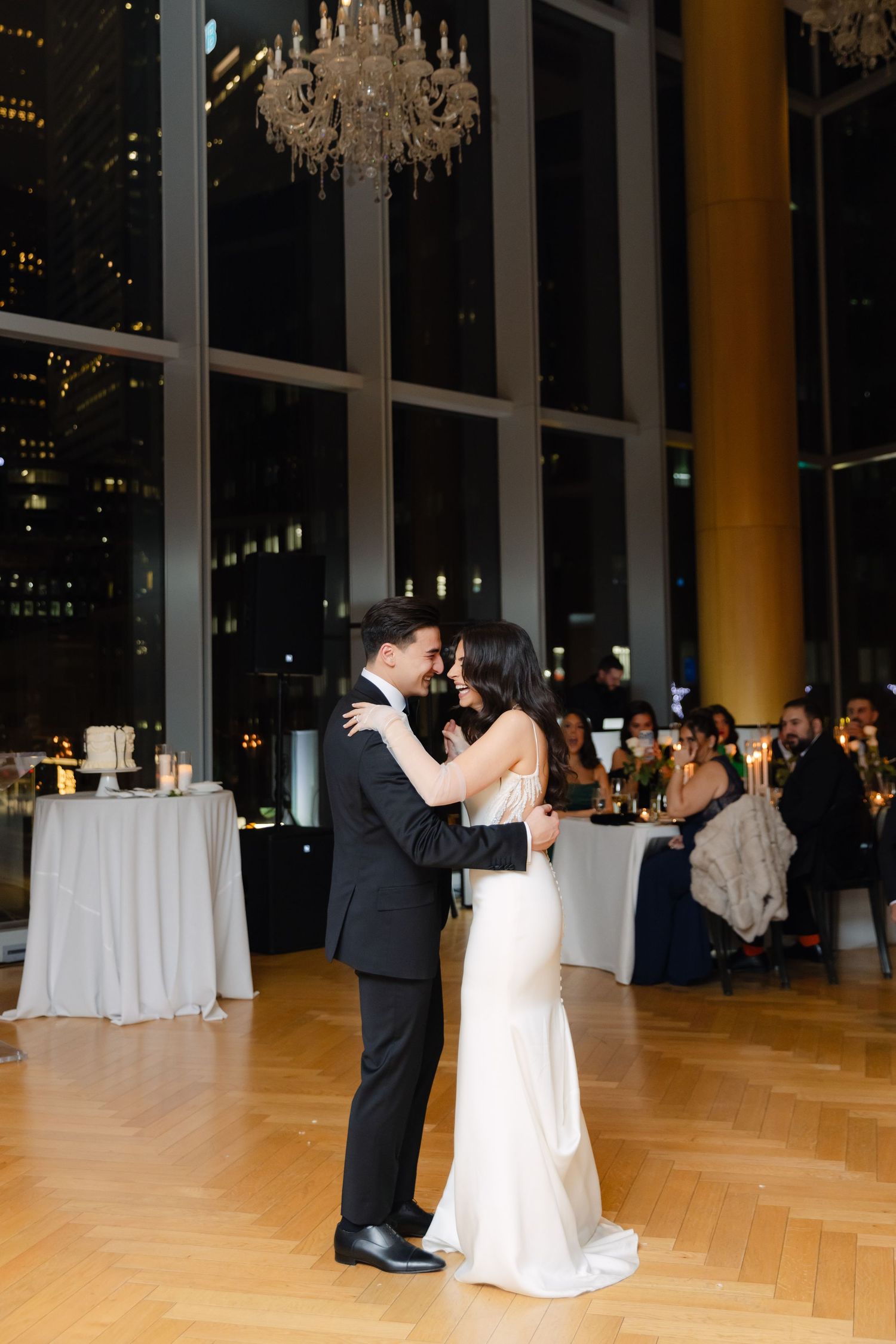 A couple embraces during their wedding dance in an elegant ballroom with city views.