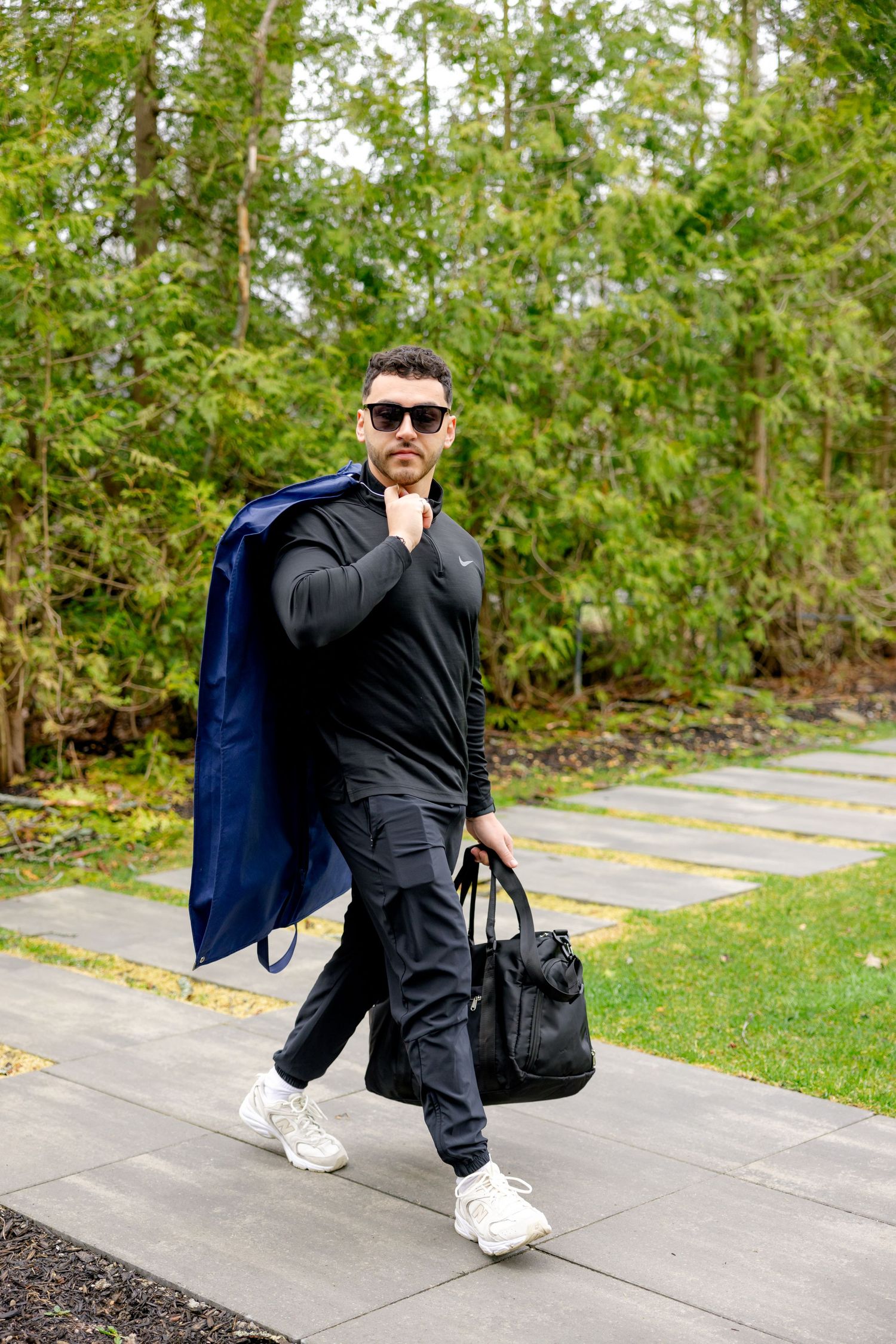 Person in all black athletic wear carrying duffle bag and coat on stone path.