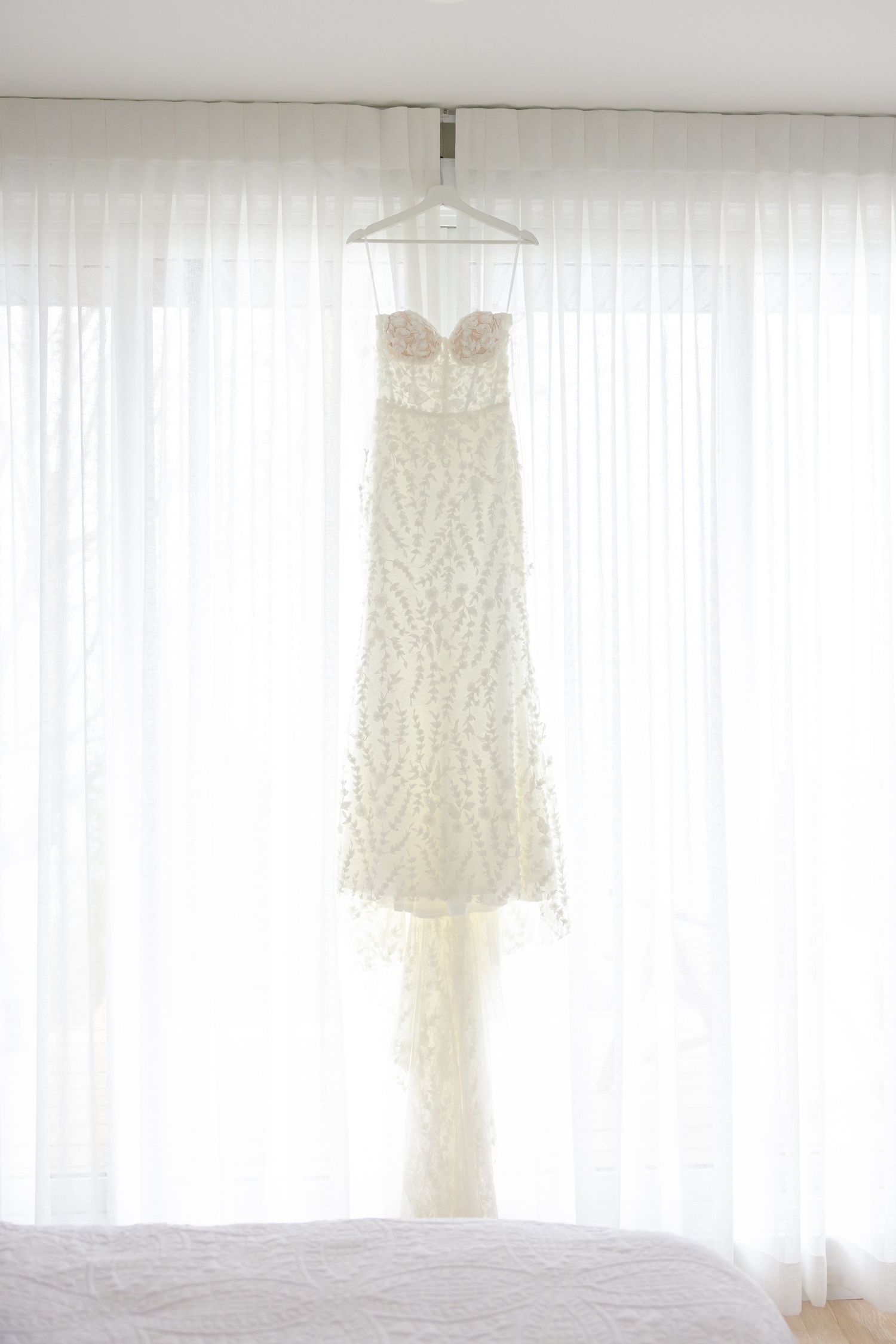 Delicate lace wedding dress hangs against sheer white curtains in natural light.