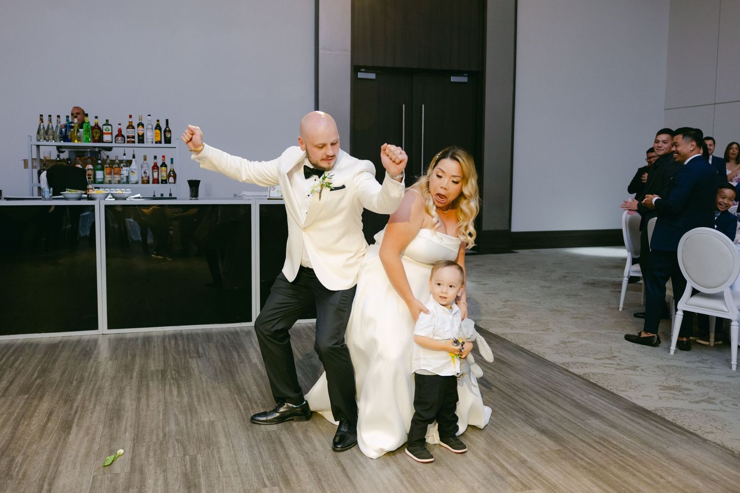 A family dances together at a wedding reception on a wooden dance floor next to a bar area.