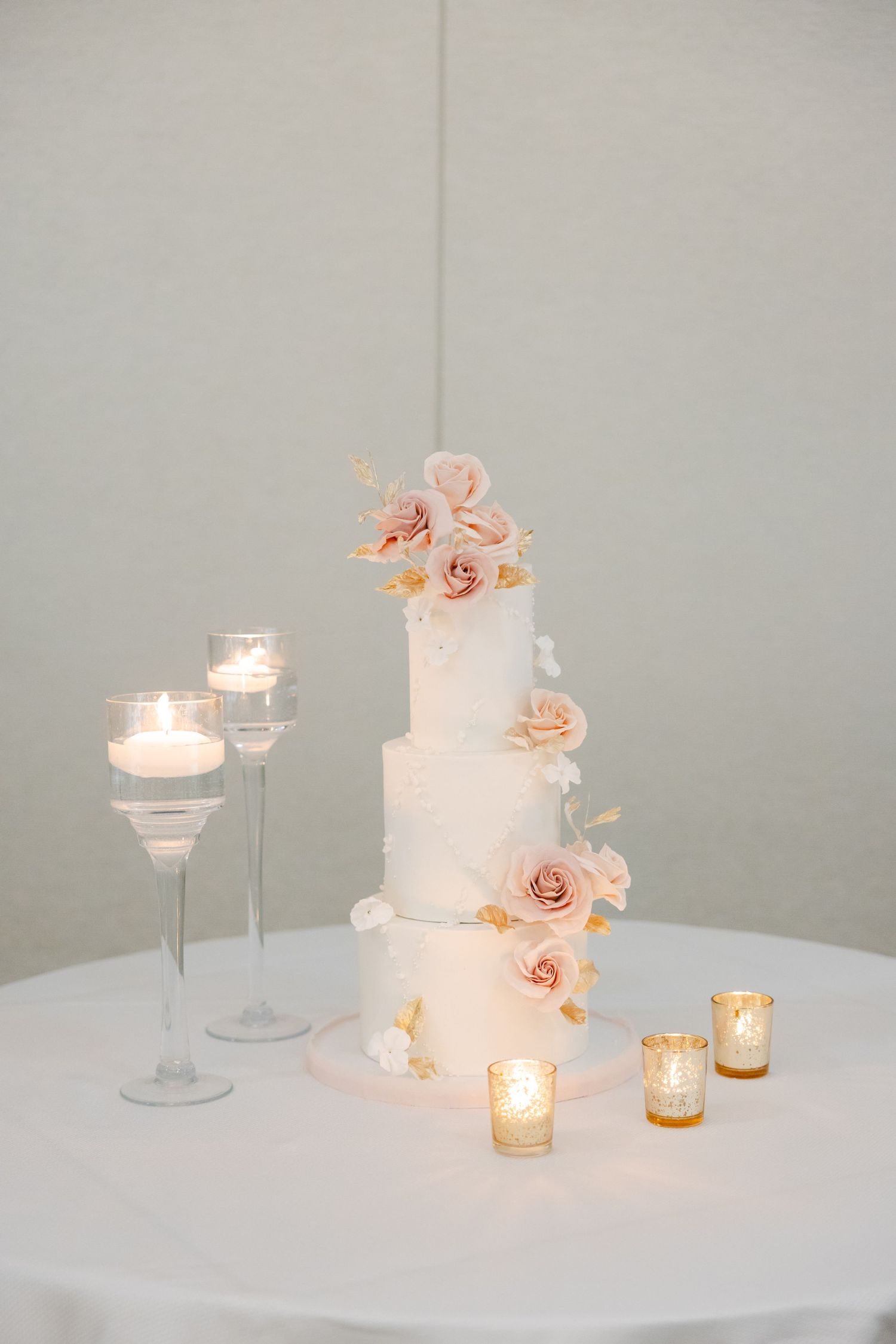 White tiered wedding cake decorated with blush roses and surrounded by glowing candlelight.