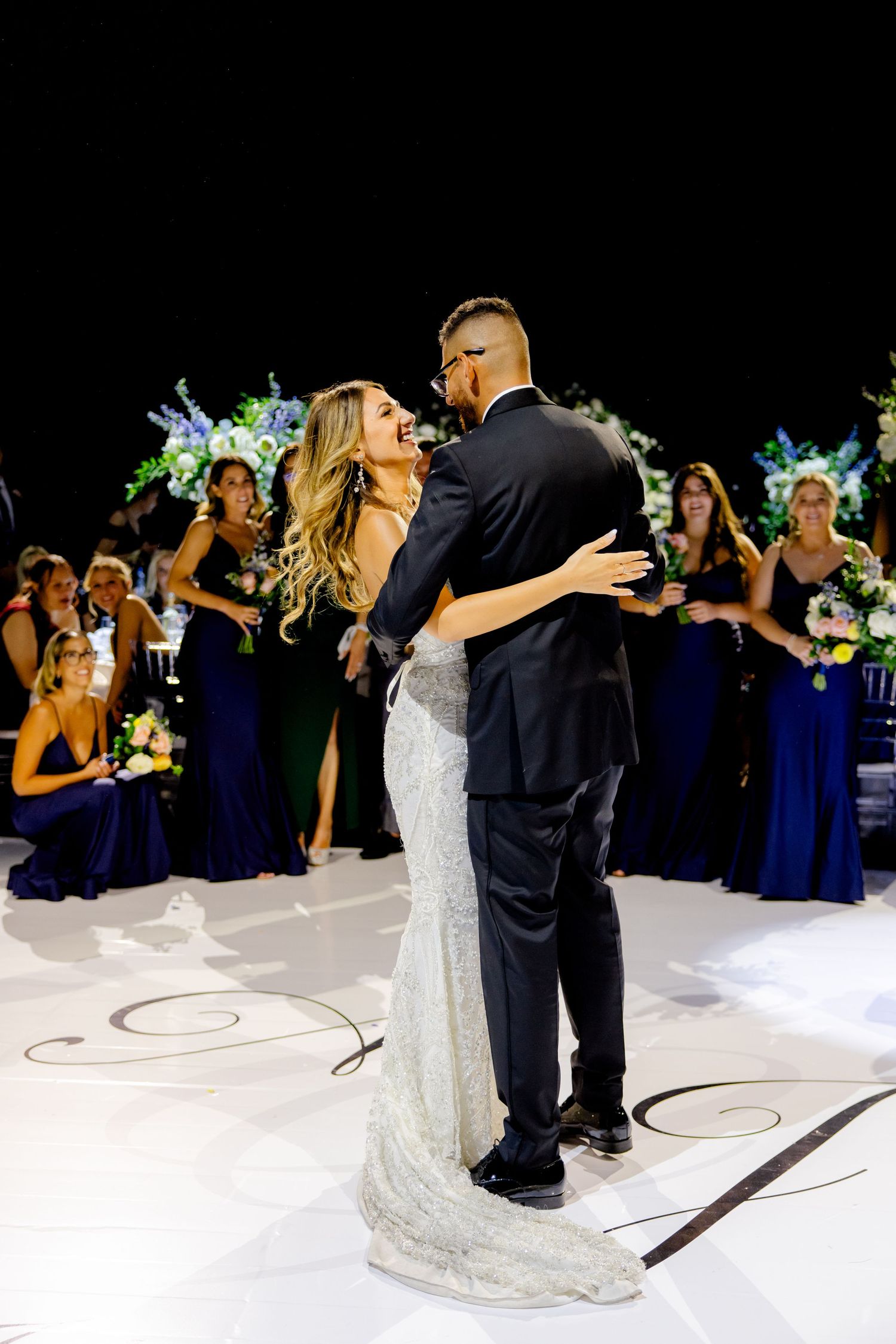 A romantic wedding dance moment captured with floral decorations and navy-clad bridesmaids in background.