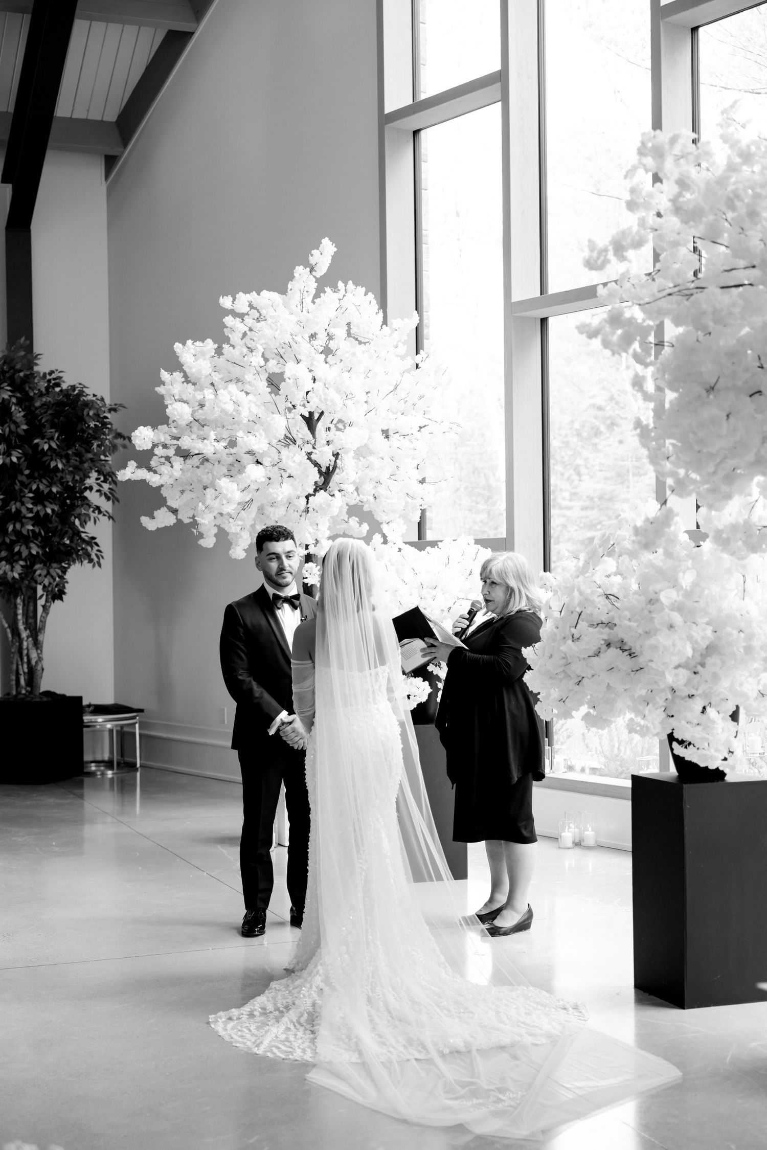 An elegant wedding ceremony setup with towering white floral arrangements and floor-to-ceiling windows.