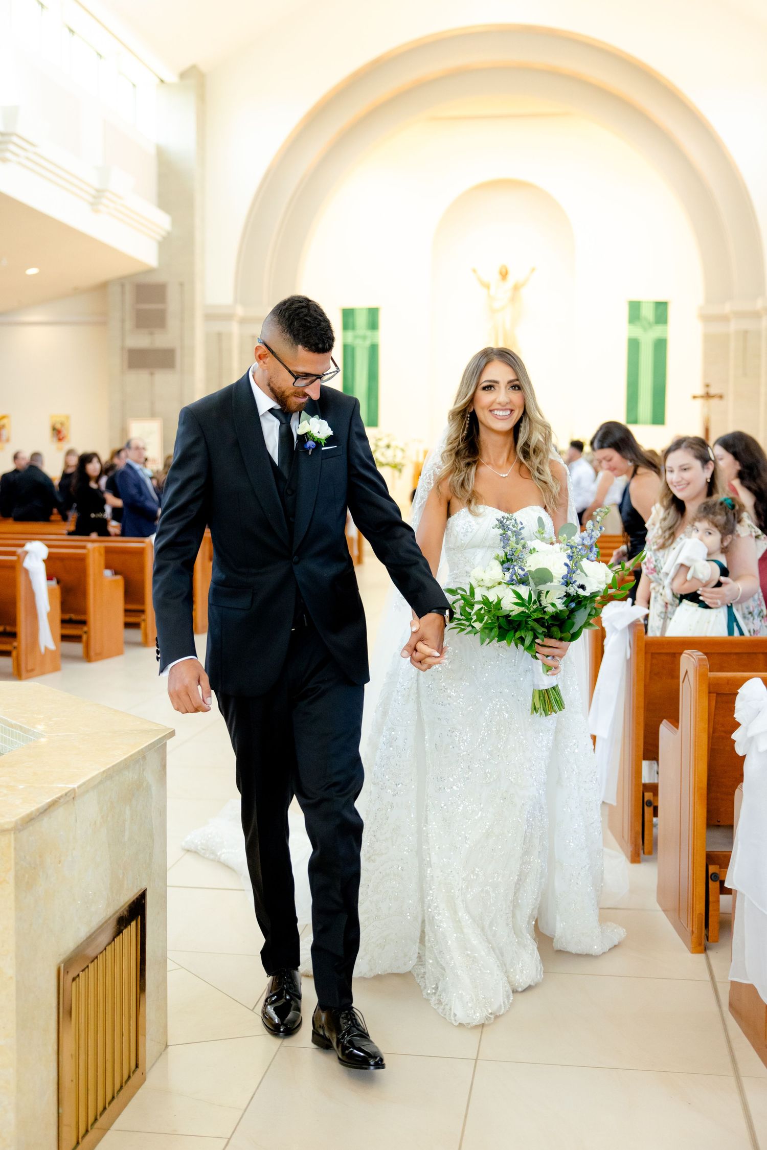 Newlyweds walk down the church aisle holding hands while wedding guests look on from wooden pews.