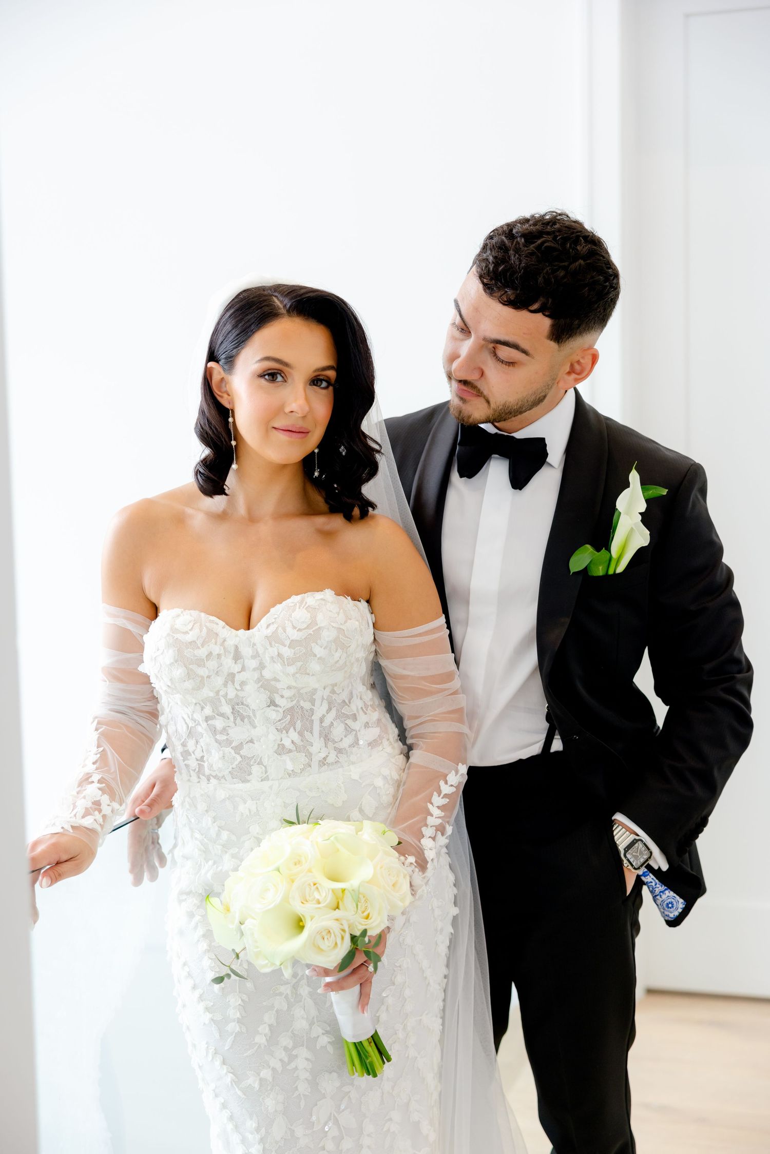 A wedding couple poses together, with the bride wearing a strapless lace gown and holding a cream-colored bouquet.
