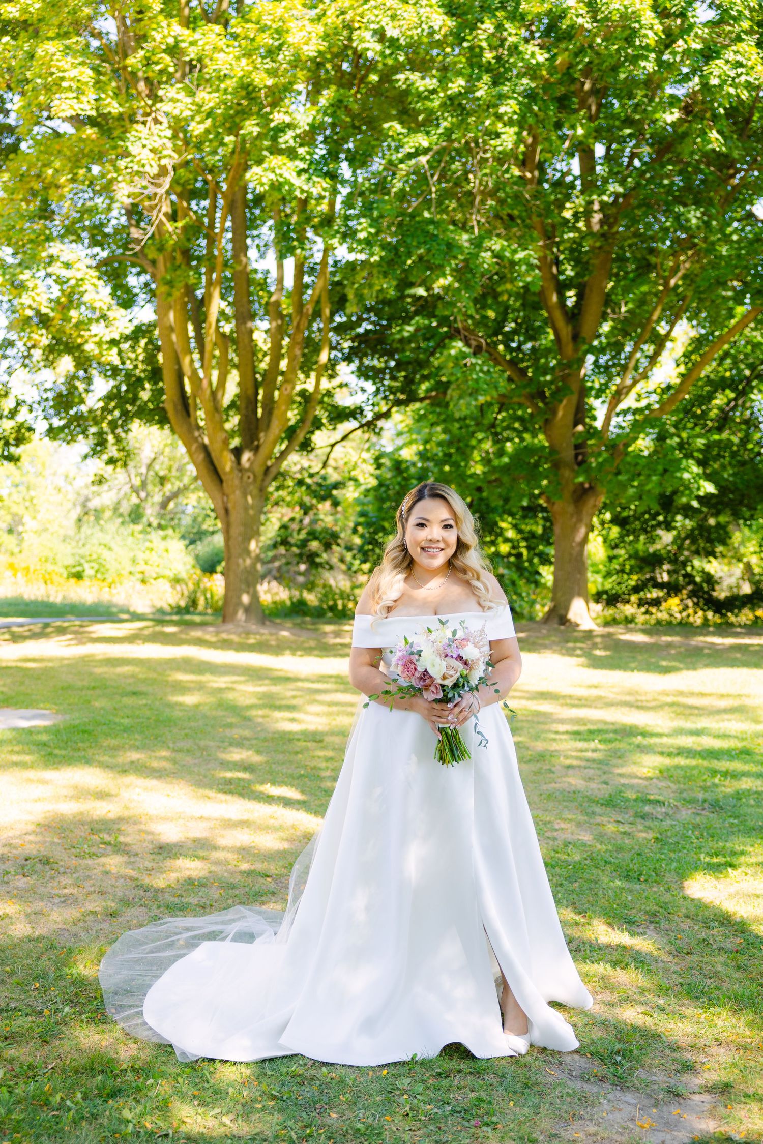 Bride in off-shoulder wedding gown holds pink and white bouquet in sunlit garden setting.
