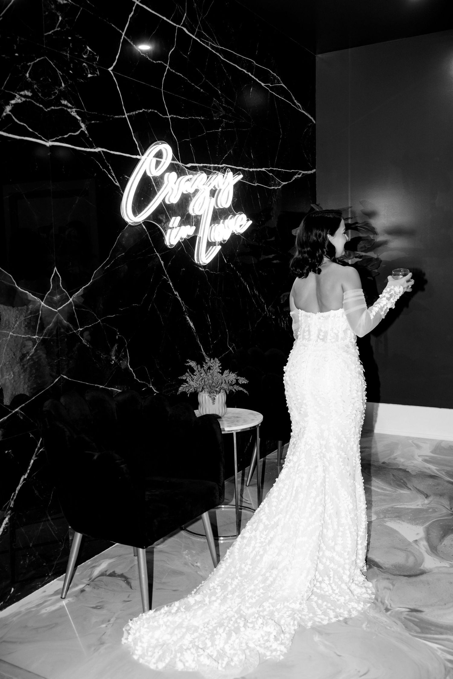 Black and white photo of bride in fitted lace gown against dark marble wall with illuminated signage.