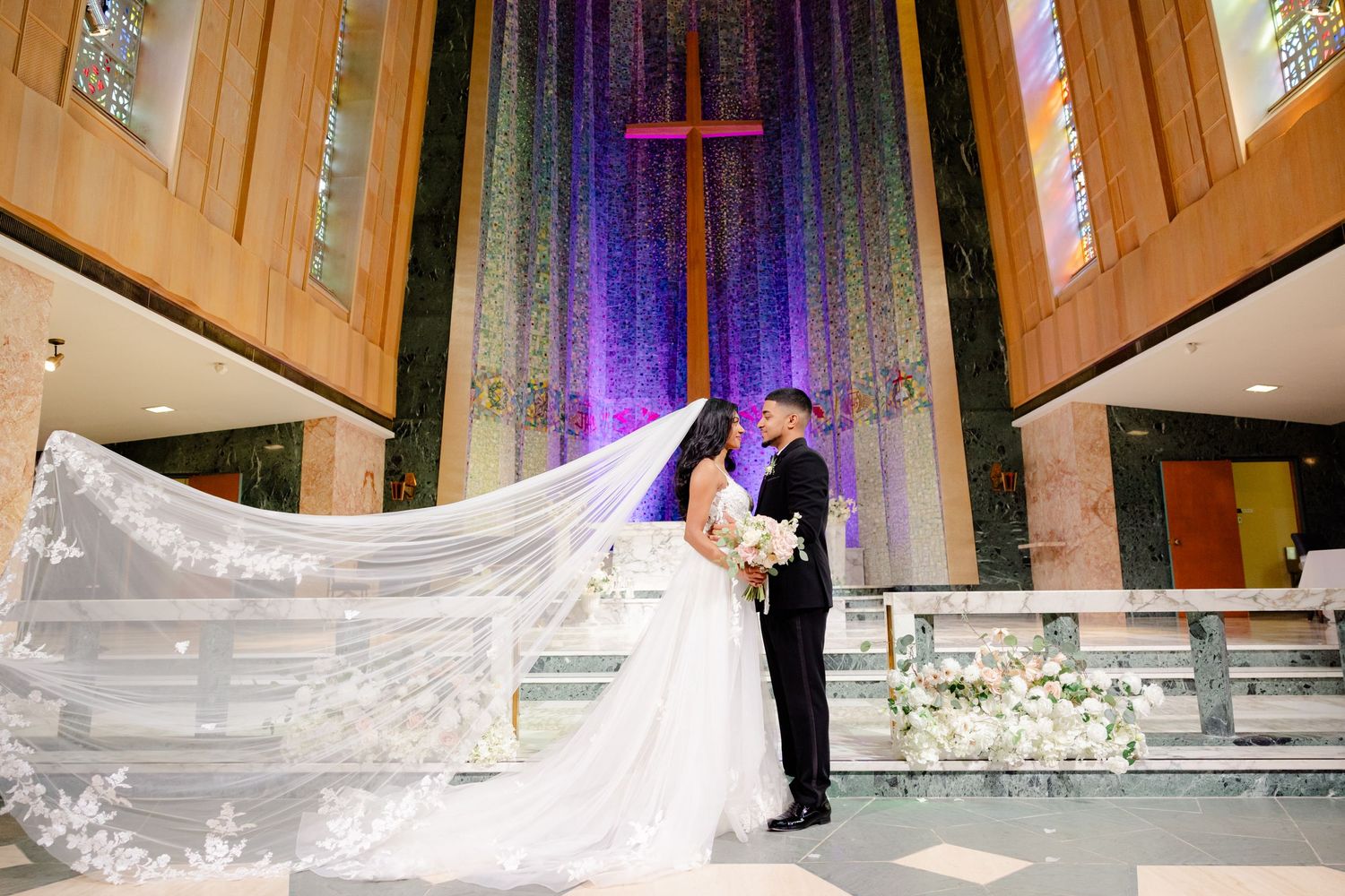 Romantic wedding moment in front of illuminated purple cross with flowing cathedral veil in church sanctuary.