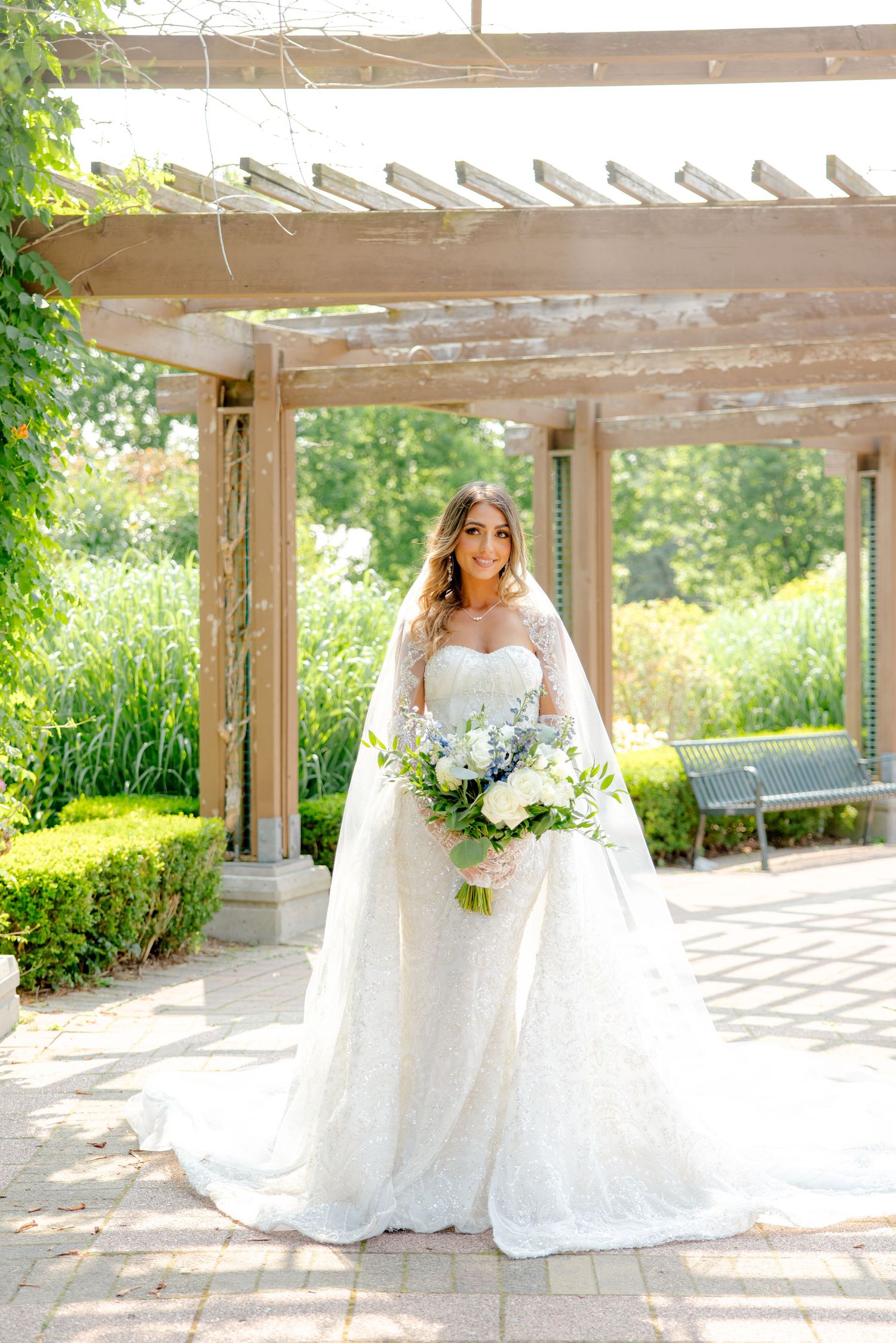 Full-length bridal portrait in sunlit garden pergola featuring detailed lace gown and floral bouquet.