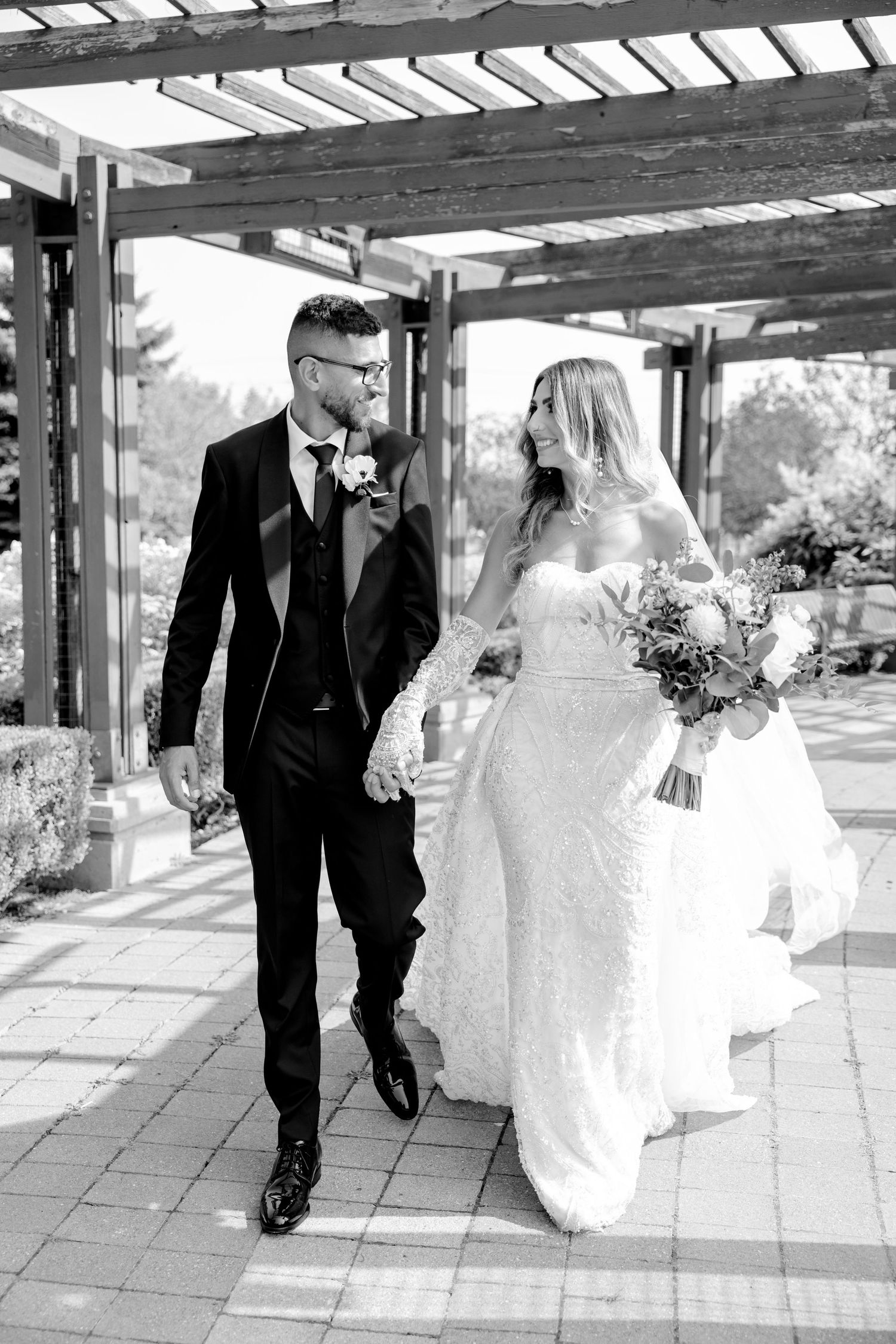 Black and white photo of couple walking together under rustic pergola.