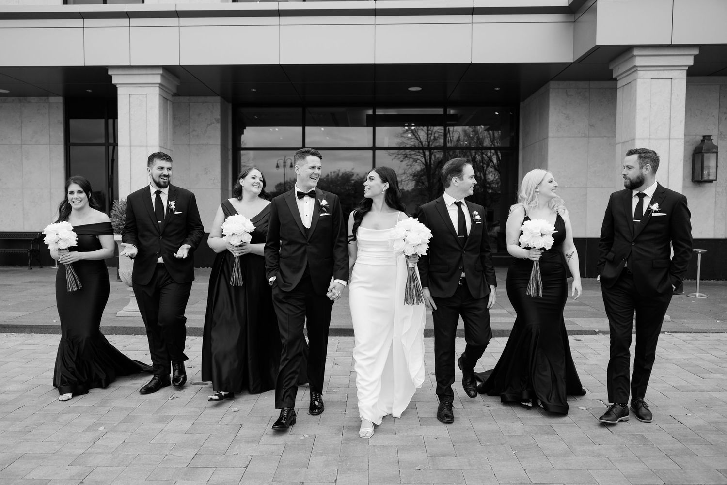 Black and white photo of wedding party walking and laughing together outside a modern building.