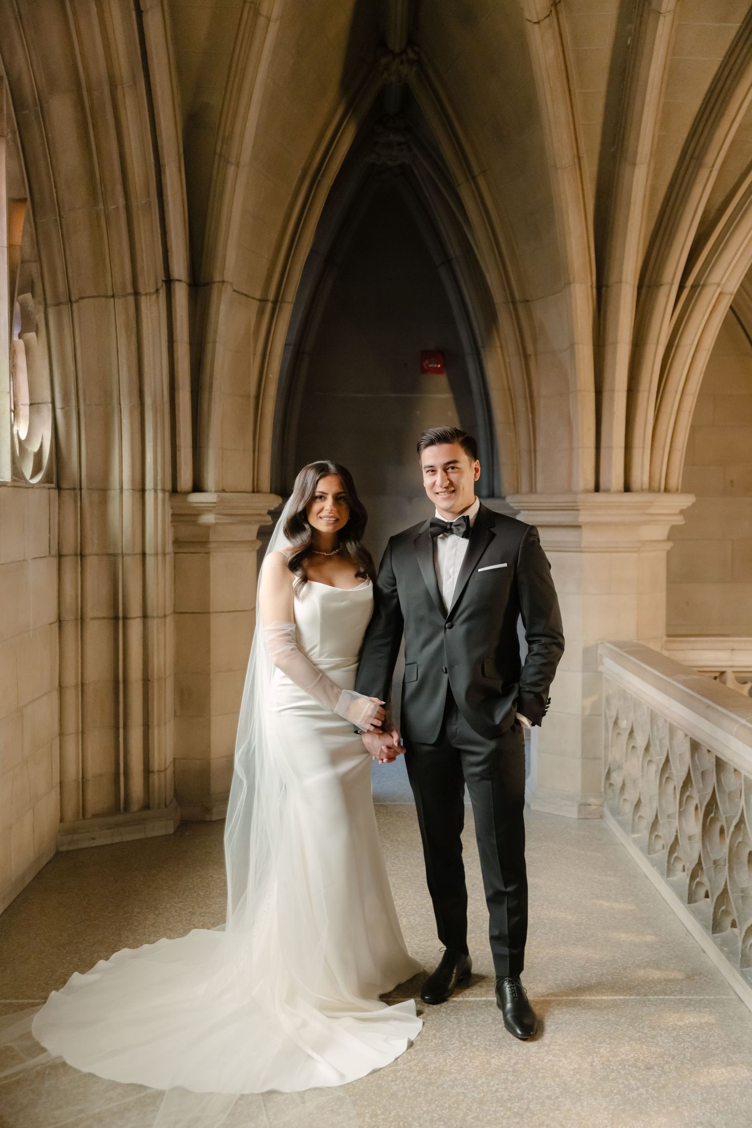 A wedding couple poses in formal attire beneath dramatic gothic cathedral arches and stone columns.
