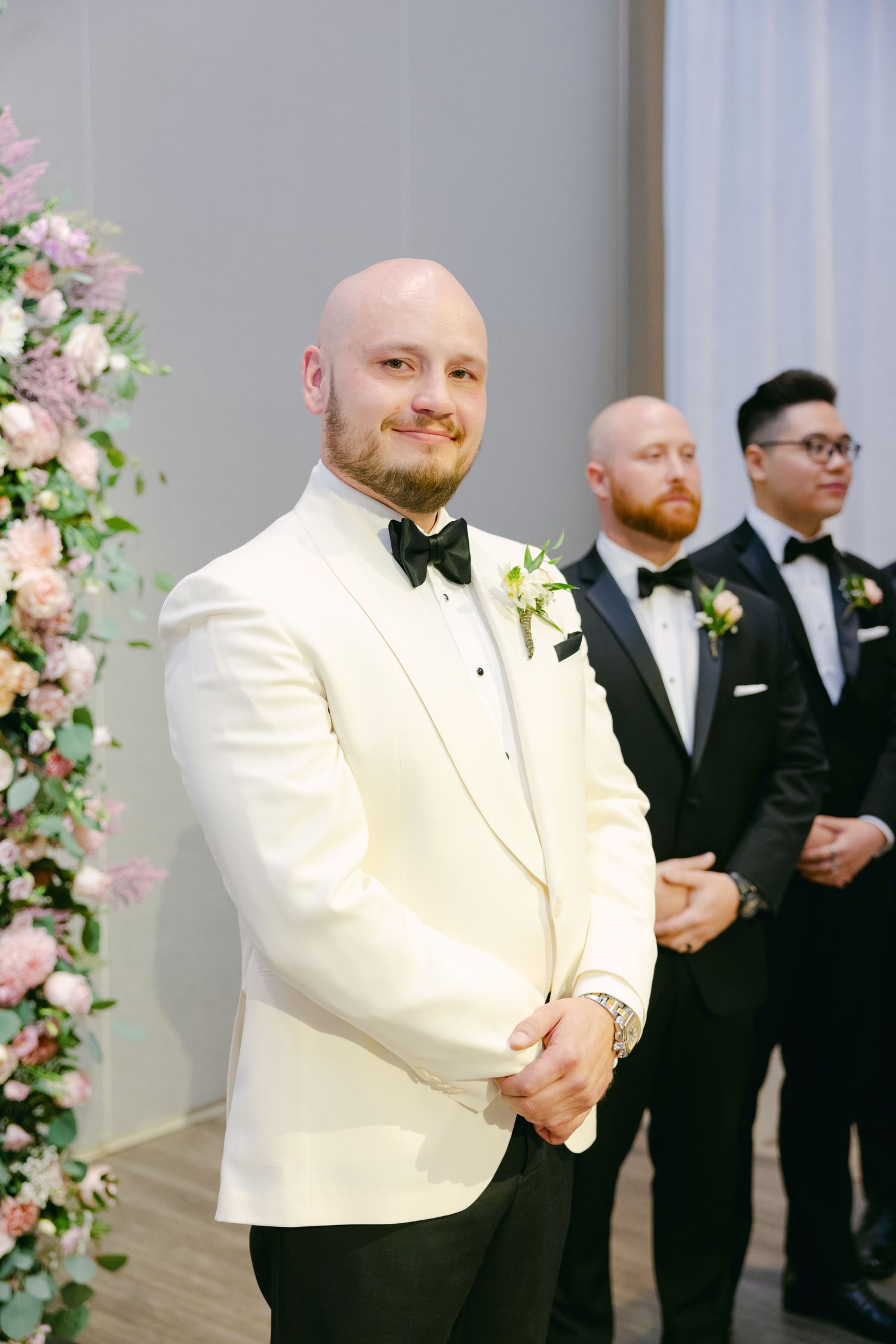 A groom in a white dinner jacket stands at the altar with groomsmen in black tuxedos.