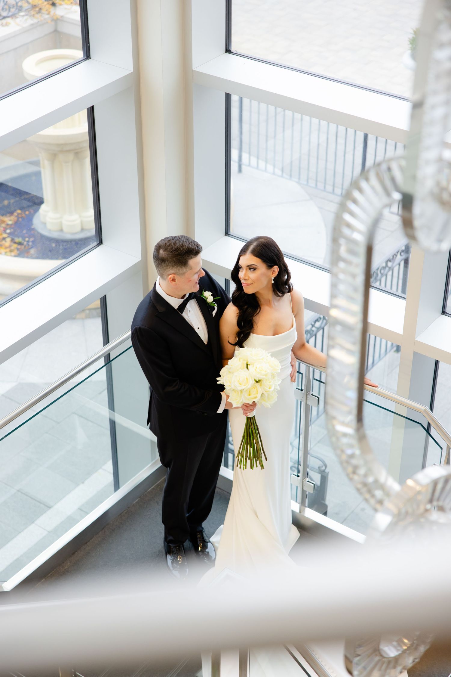 A couple in wedding attire share an intimate moment in a modern stairwell with large windows and a white bouquet.