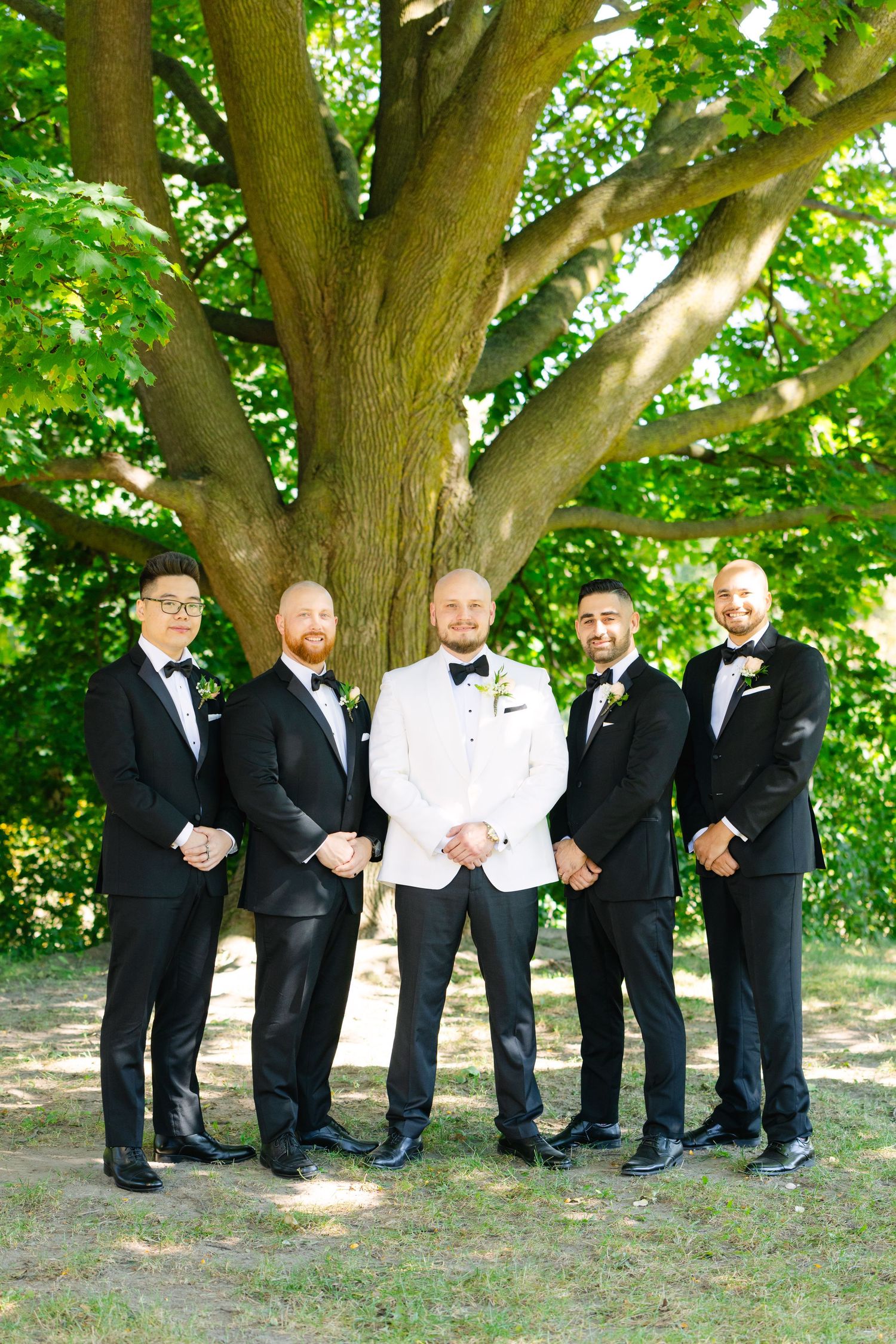 Groomsmen in formal black suits and white jacket pose together under a large tree at an outdoor wedding.