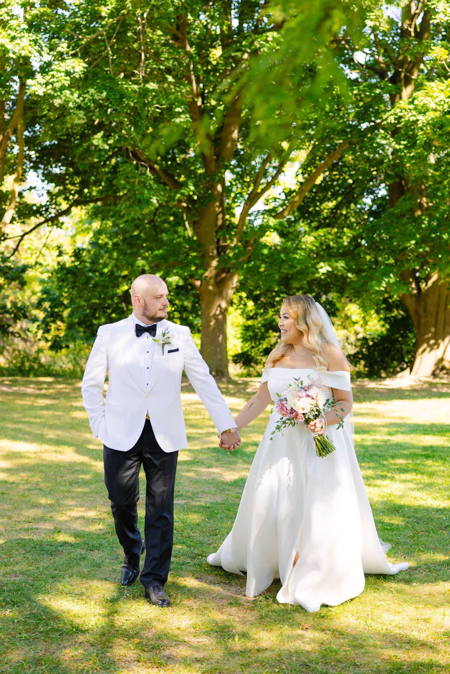 Wedding couple strolls together on sunny lawn with green trees in background.